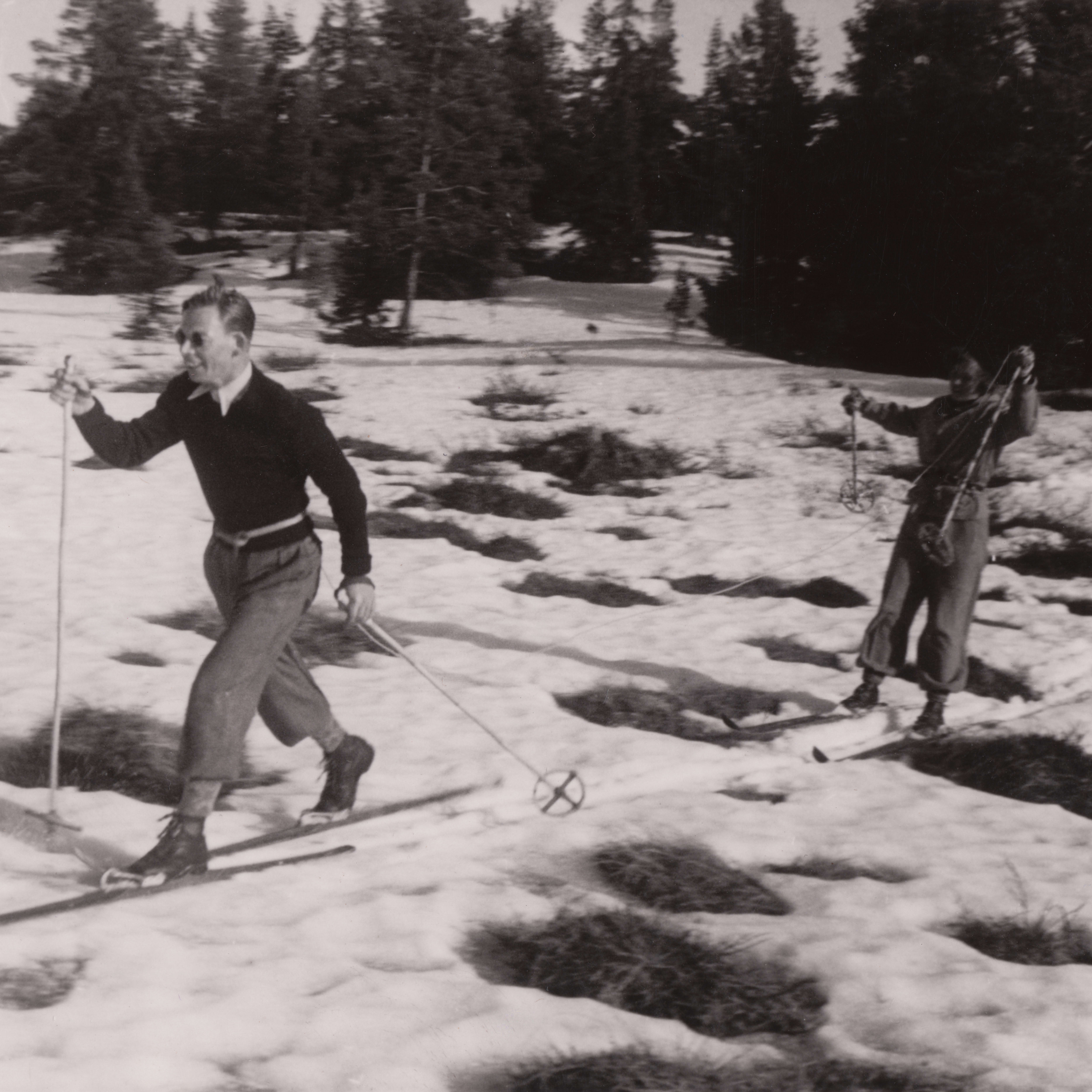 Two skiers in Norrøna clothing traverse a snowy, grassy field toward trees in this black-and-white vintage photo.