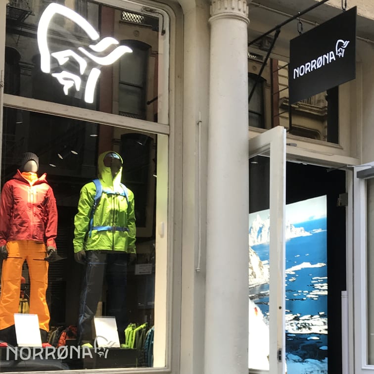 Norrøna storefront with mannequins wearing Norrøna jackets and pants. A sign above the door reads "Norrøna".  A photo of mountains and ocean is visible through the open door.  
