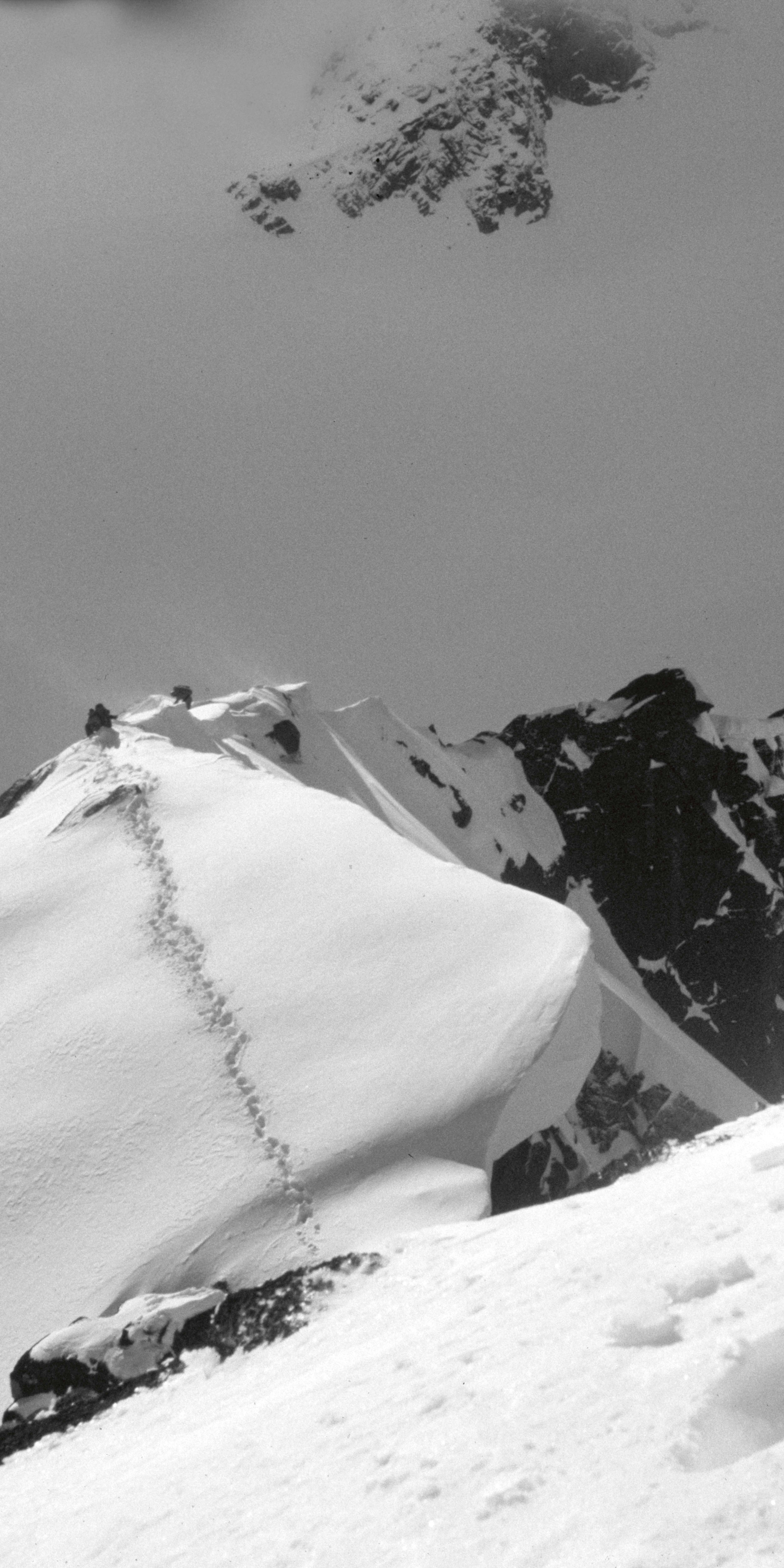 A black and white image of a snowy mountain peak with a trail of footprints leading up to the summit.  The peak is partially obscured by clouds. 
