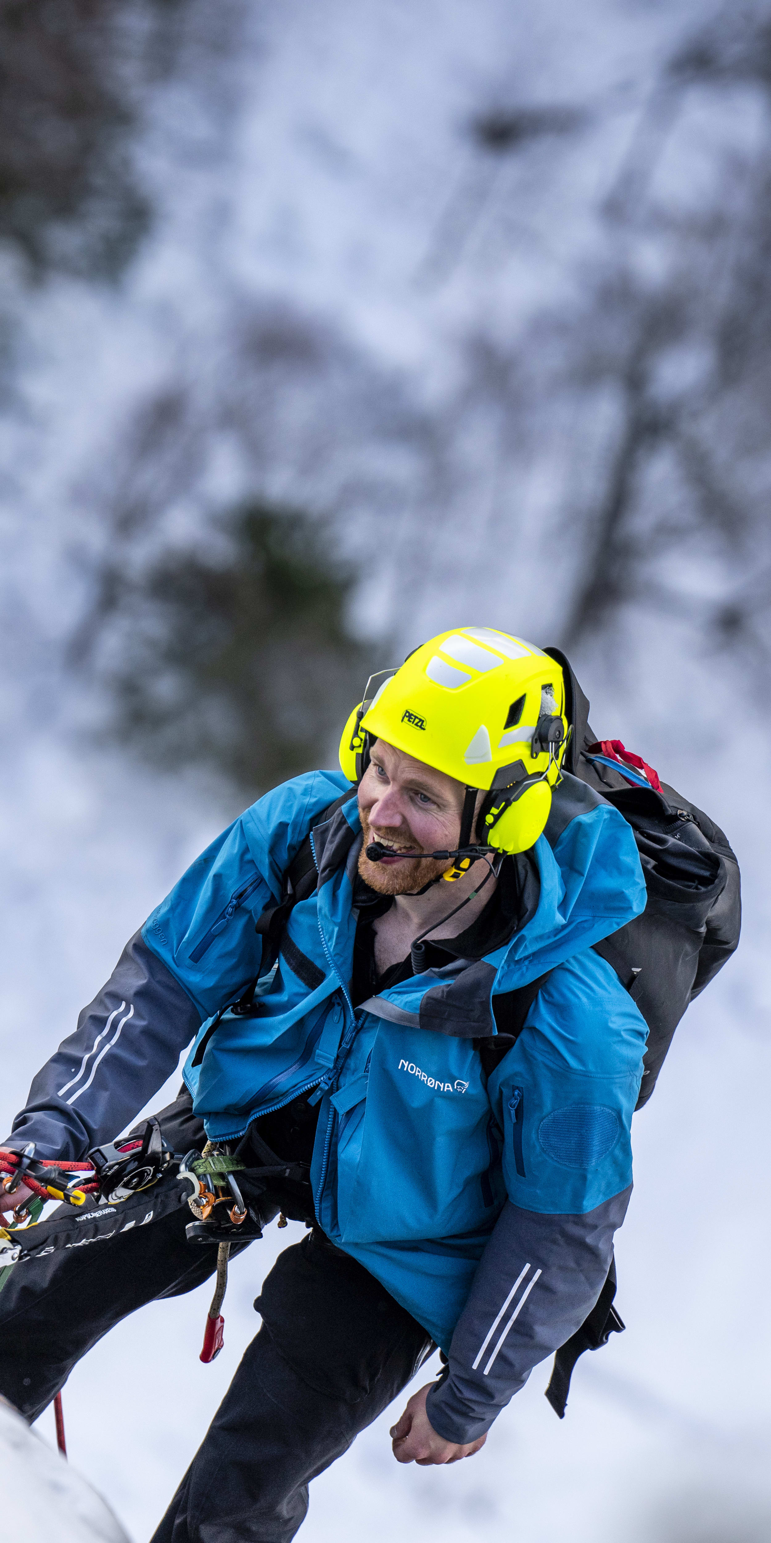 A man wearing a Norrøna jacket and a helmet climbs an icy wall. A red rope secures him to the mountain.