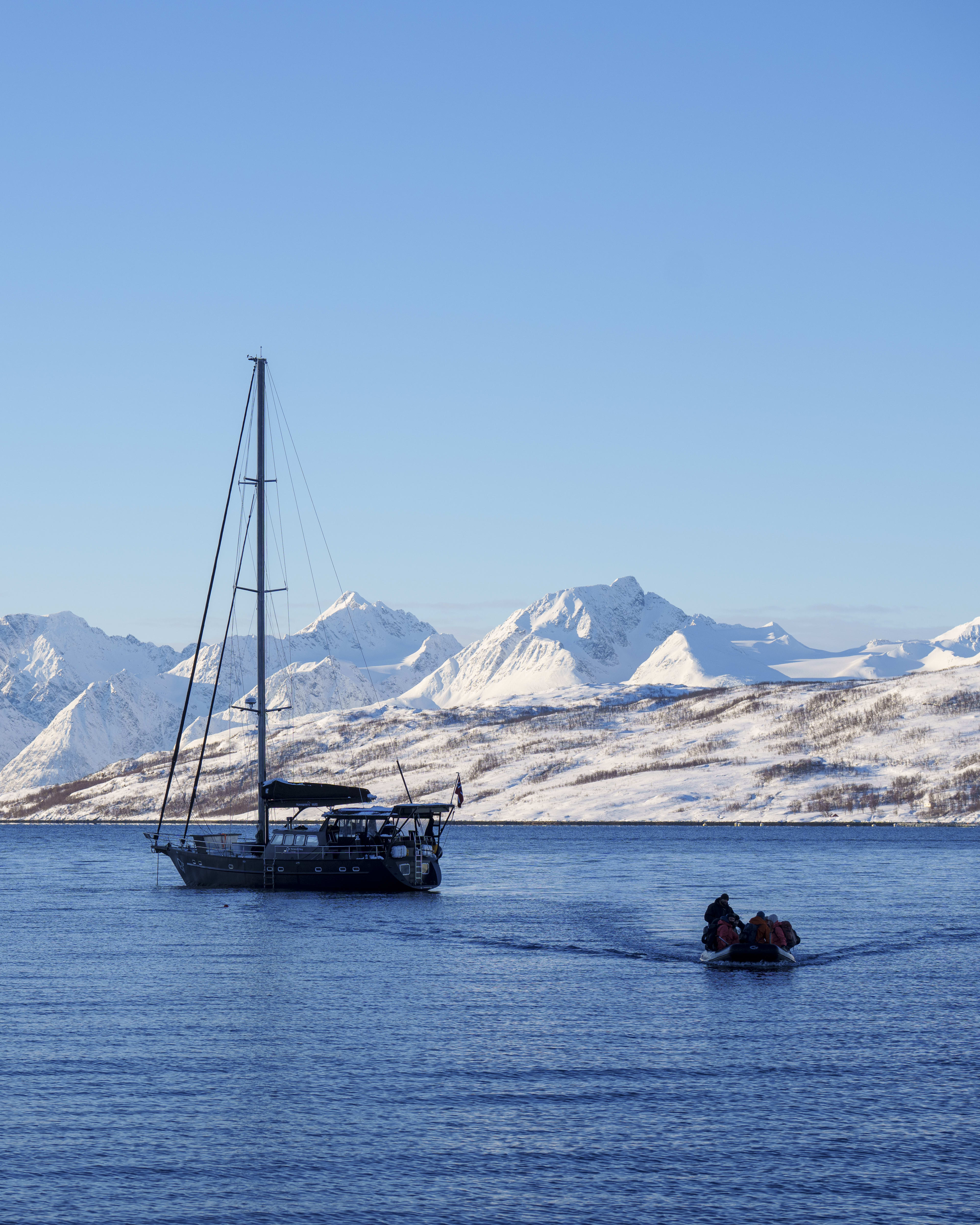 A sailboat sits on blue water with snow-covered mountains behind. A small boat approaches it, the people on it are wearing Norrøna gear.