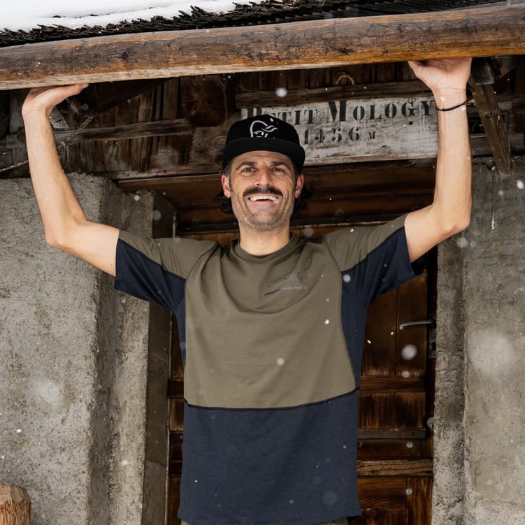 A smiling man stands under a snowy roof, wearing a black Norrøna cap and a two-tone t-shirt. He holds the roof beam with both hands.