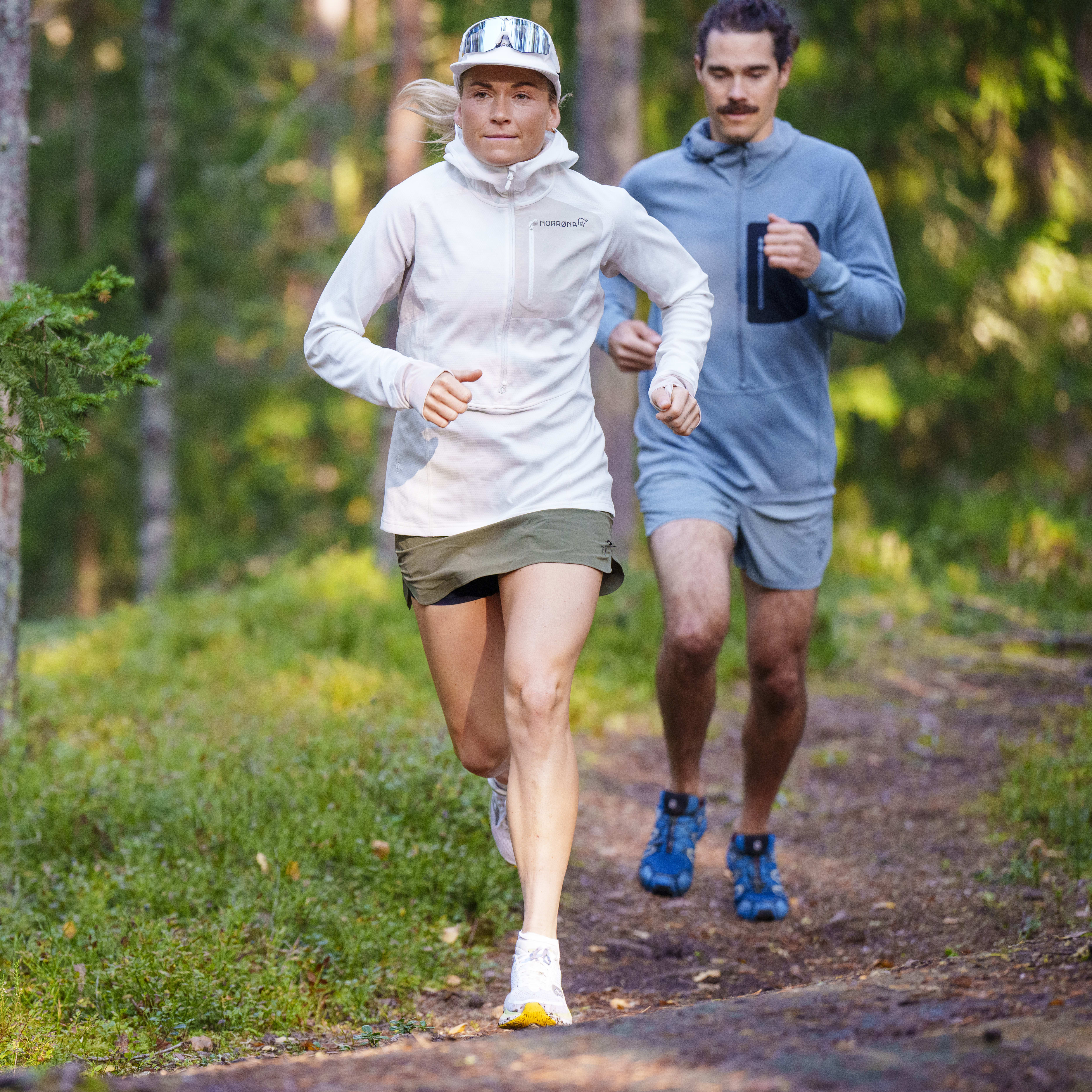 Two people run on a trail in a forest. A woman wearing a Norrøna jacket leads the way.  
