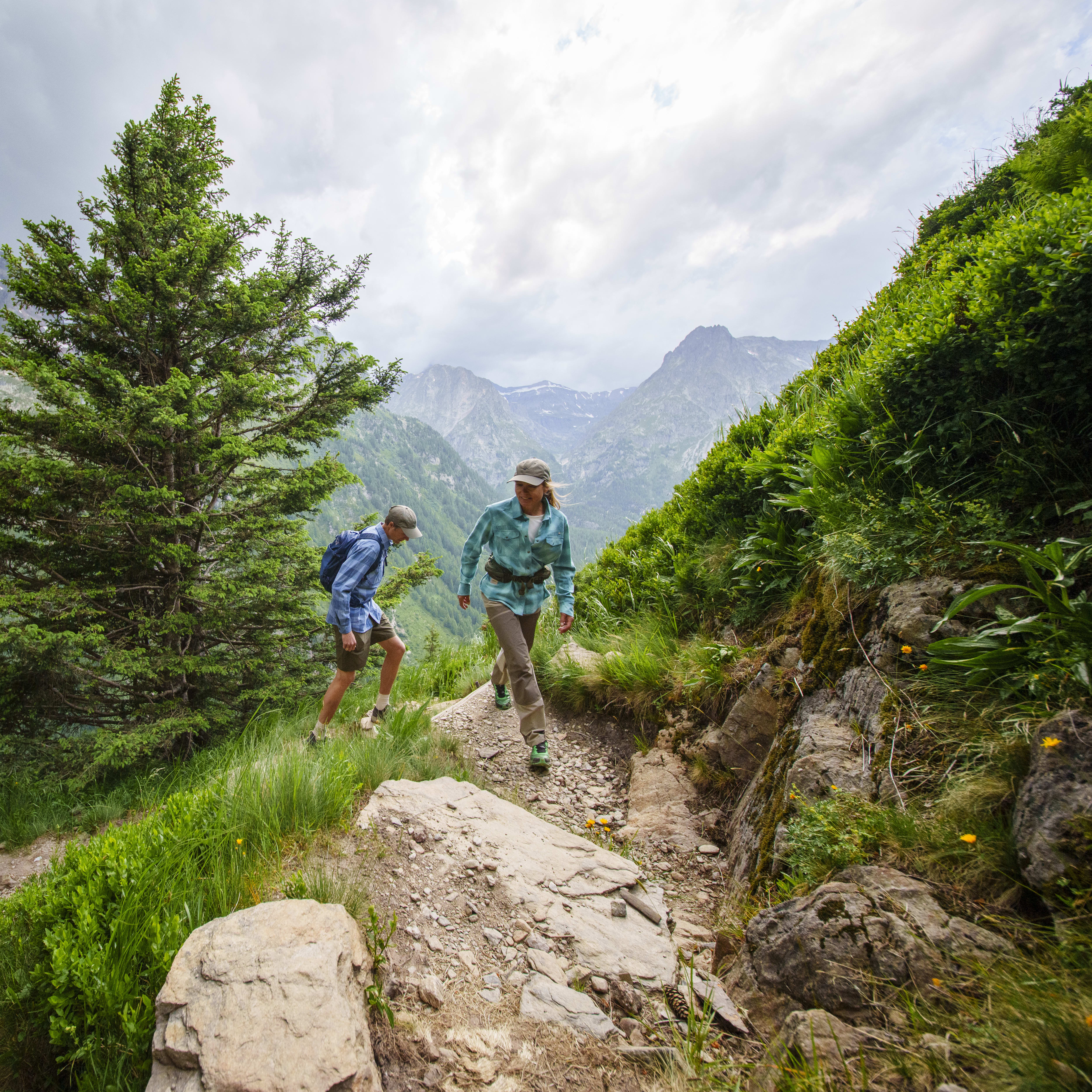 Two hikers on a mountain trail; the woman wears a Norrøna shirt and pants. Mountains and green vegetation fill the background.