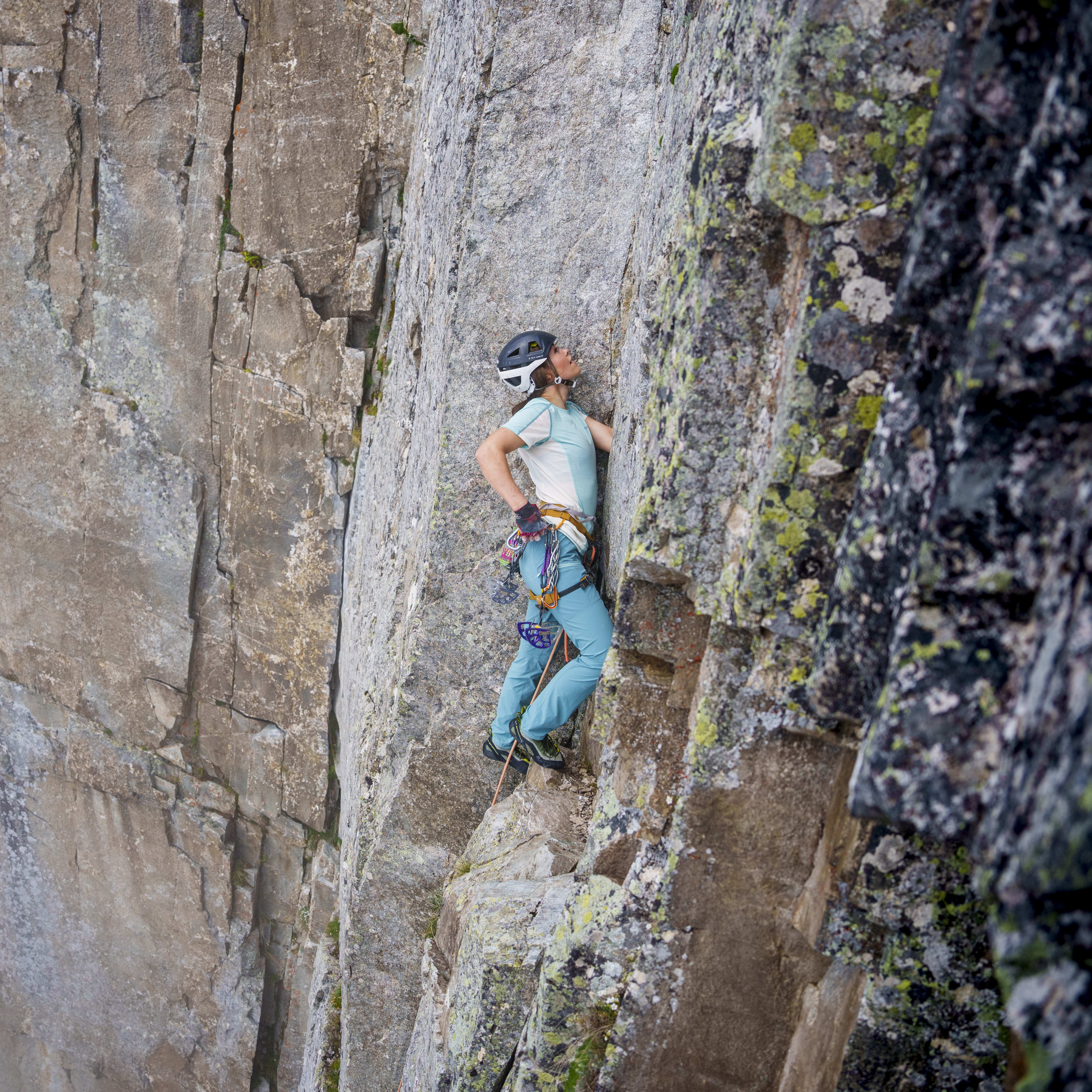 A climber in Norrøna pants scales a steep, gray rock face with climbing gear. They are wearing a helmet and looking upwards.