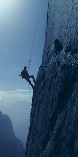 Silhouette of a climber rappelling down a steep rock face with a mountain range in the background.