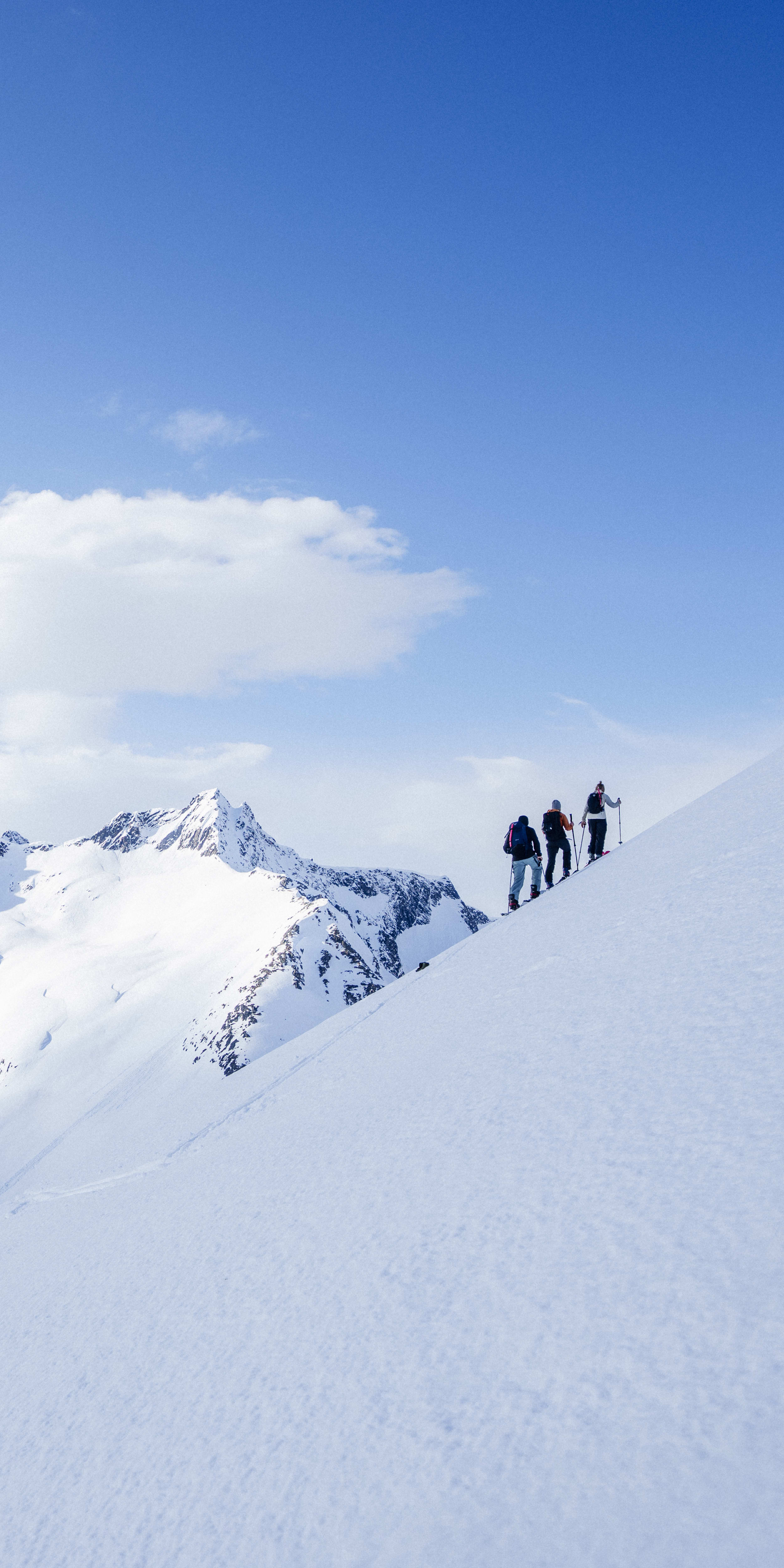 Three people hiking on a snowy mountain ridge. The sky is blue with white clouds.