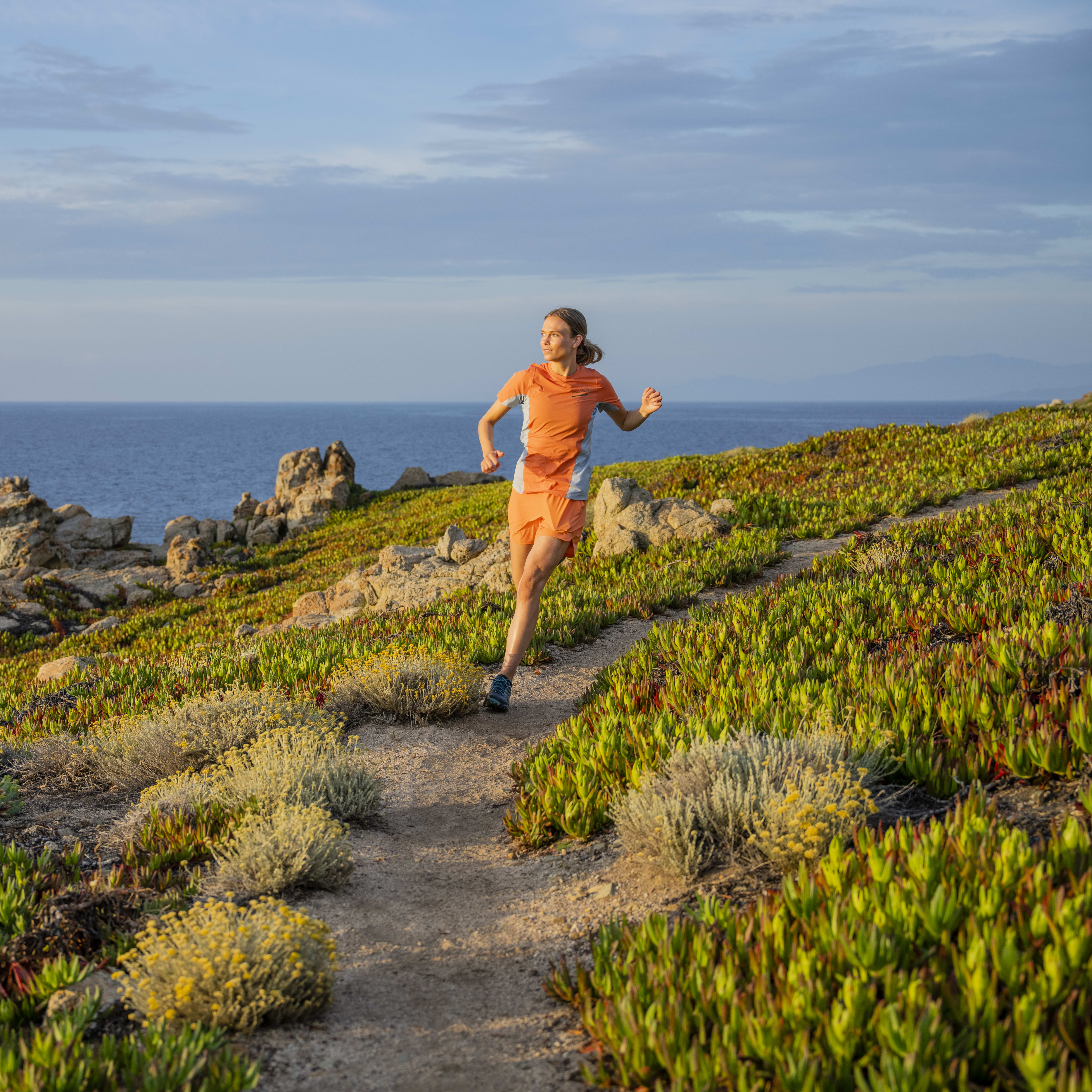 A woman in Norrøna clothing runs along a path on a cliff overlooking the ocean. 
