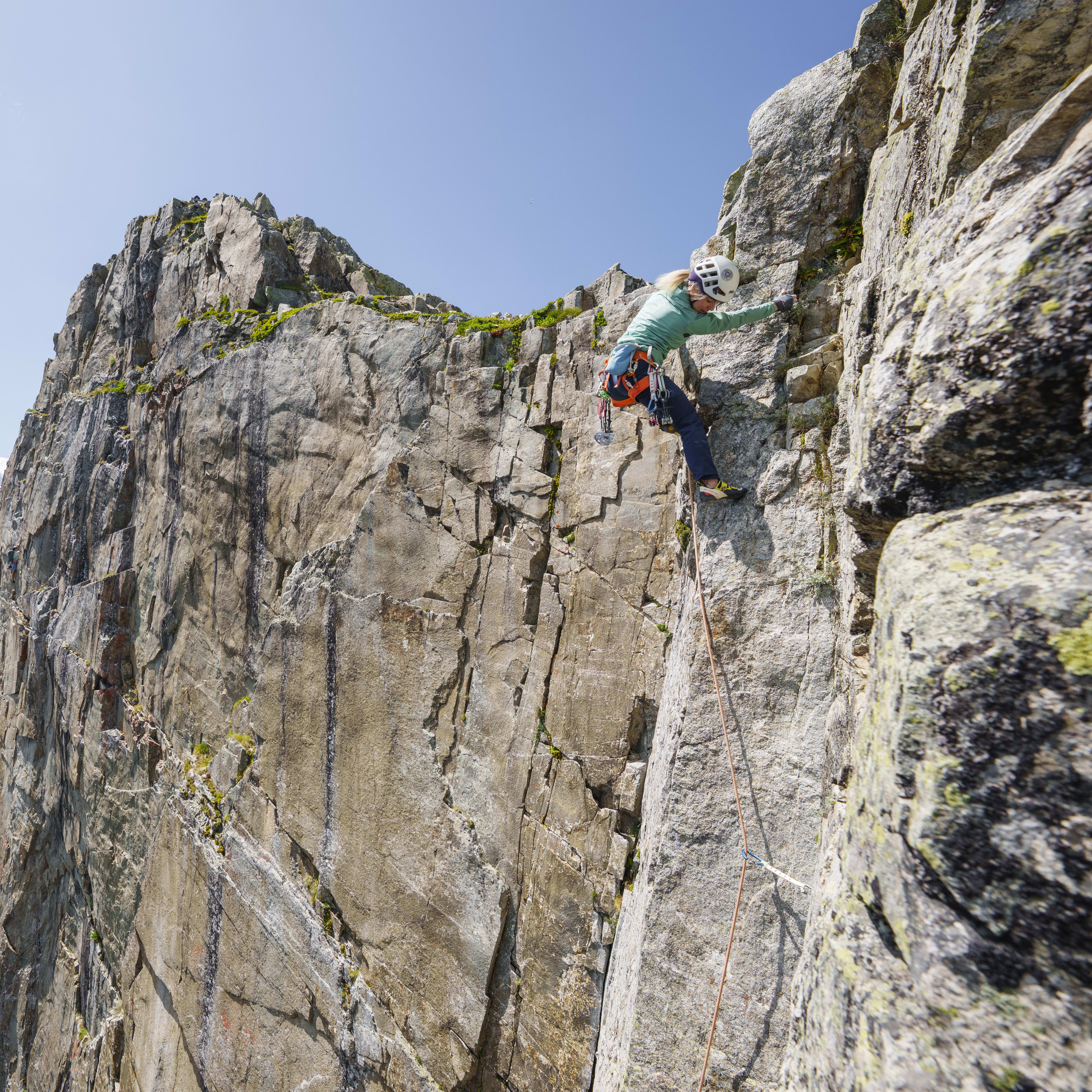 A climber scales a rock face with snow-capped mountains in the background. The climber wears a helmet and a Norrøna shirt.