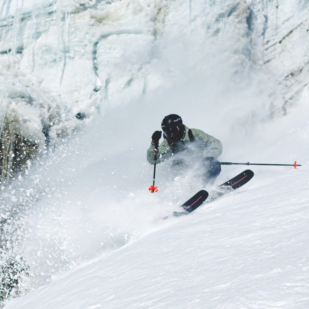A skier in a Norrøna jacket and helmet carving through fresh powder with their skis. 
