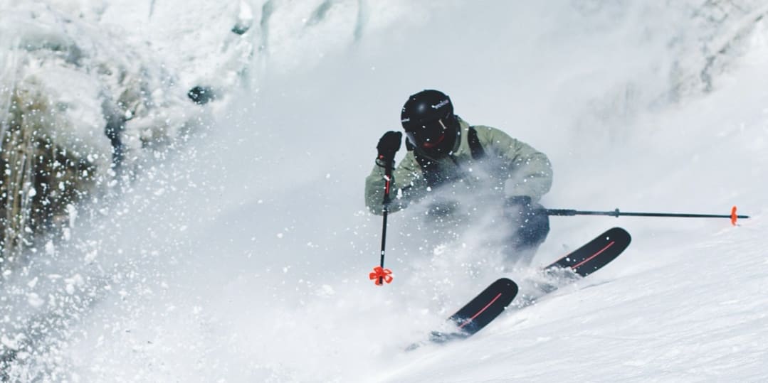A skier in a Norrøna jacket and helmet carving through fresh powder with their skis. 
