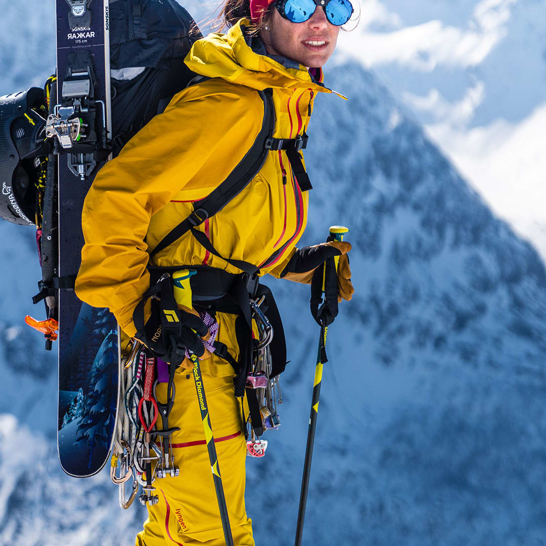 A skier in yellow Norrøna jacket and pants, with skis on their back, stands smiling against a snowy mountain backdrop.
