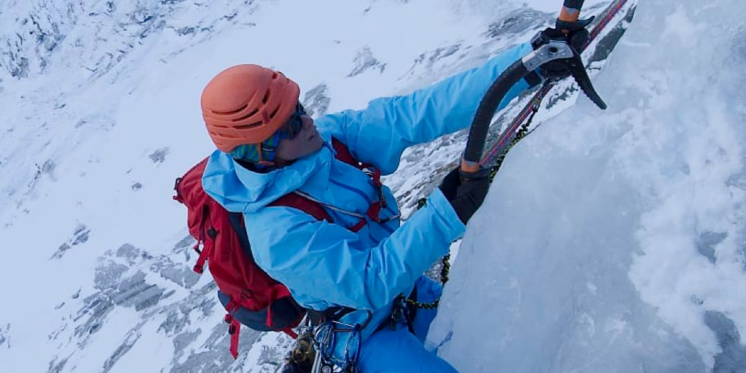 Eva Eskilsson ice climbing a steep, icy slope. She wears a helmet, sunglasses, blue Norrøna jacket and pants, a red backpack and uses ice axes.
