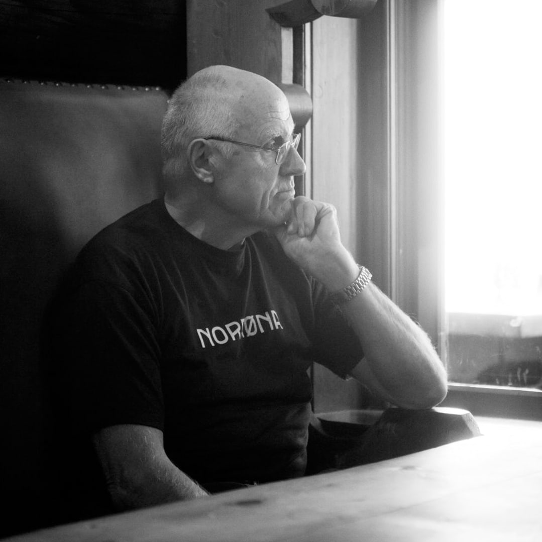 A man wearing a Norrøna shirt sits at a table, looking thoughtfully out a window.