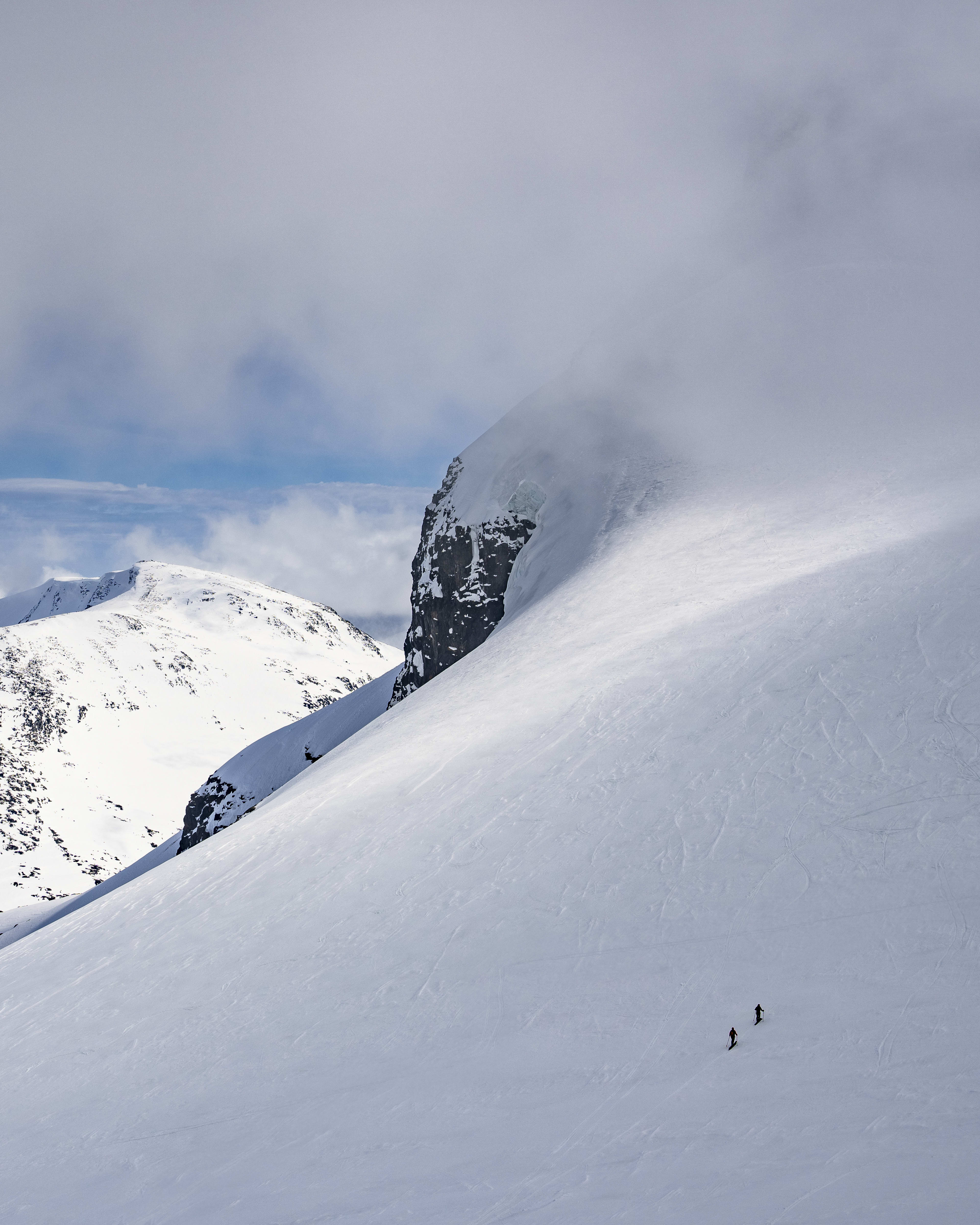 Snowy mountain landscape with distant peaks under a partly cloudy sky. Two tiny figures ski down a large slope.