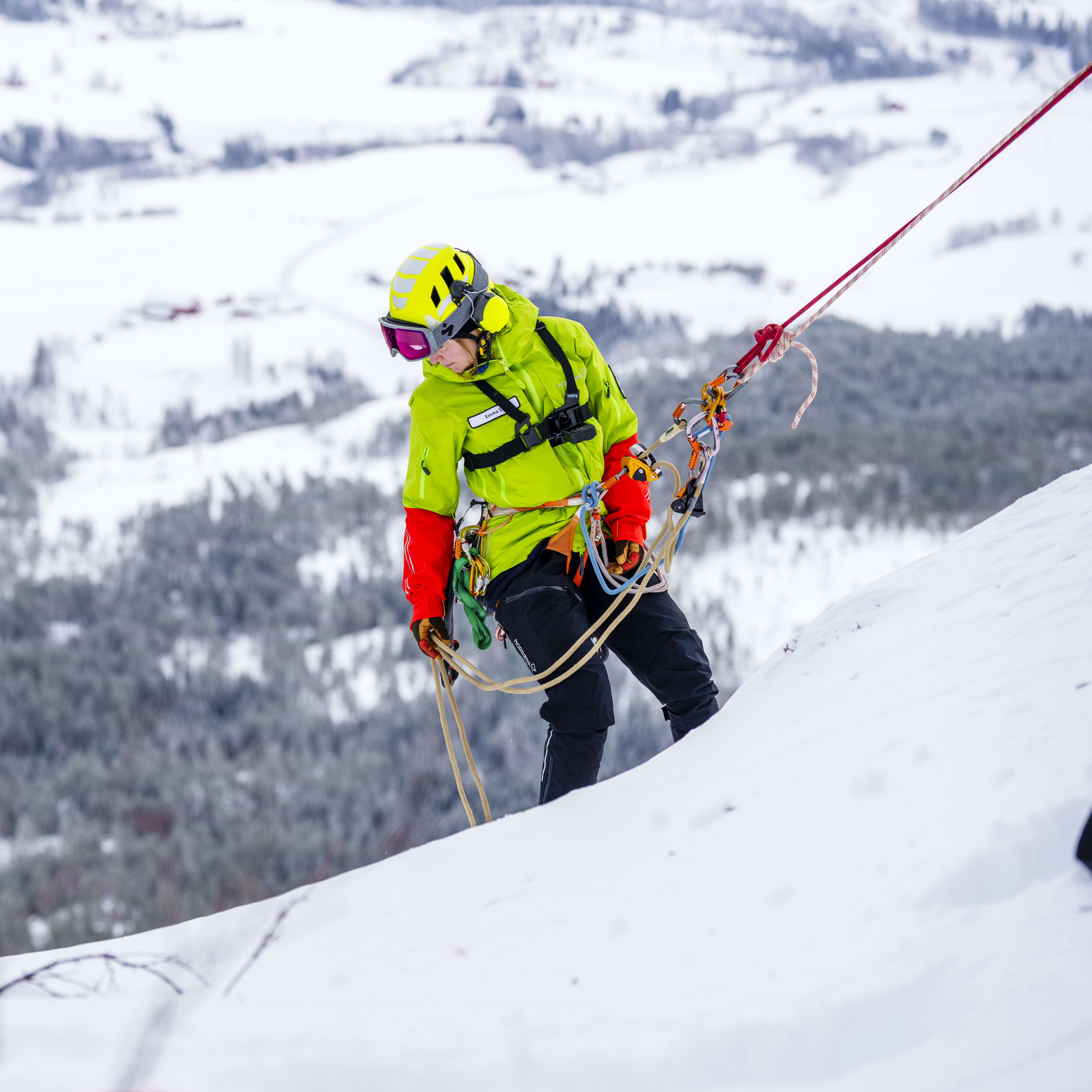 A person wearing a Norrøna jacket and a helmet is rappelling down a snow-covered mountain.  