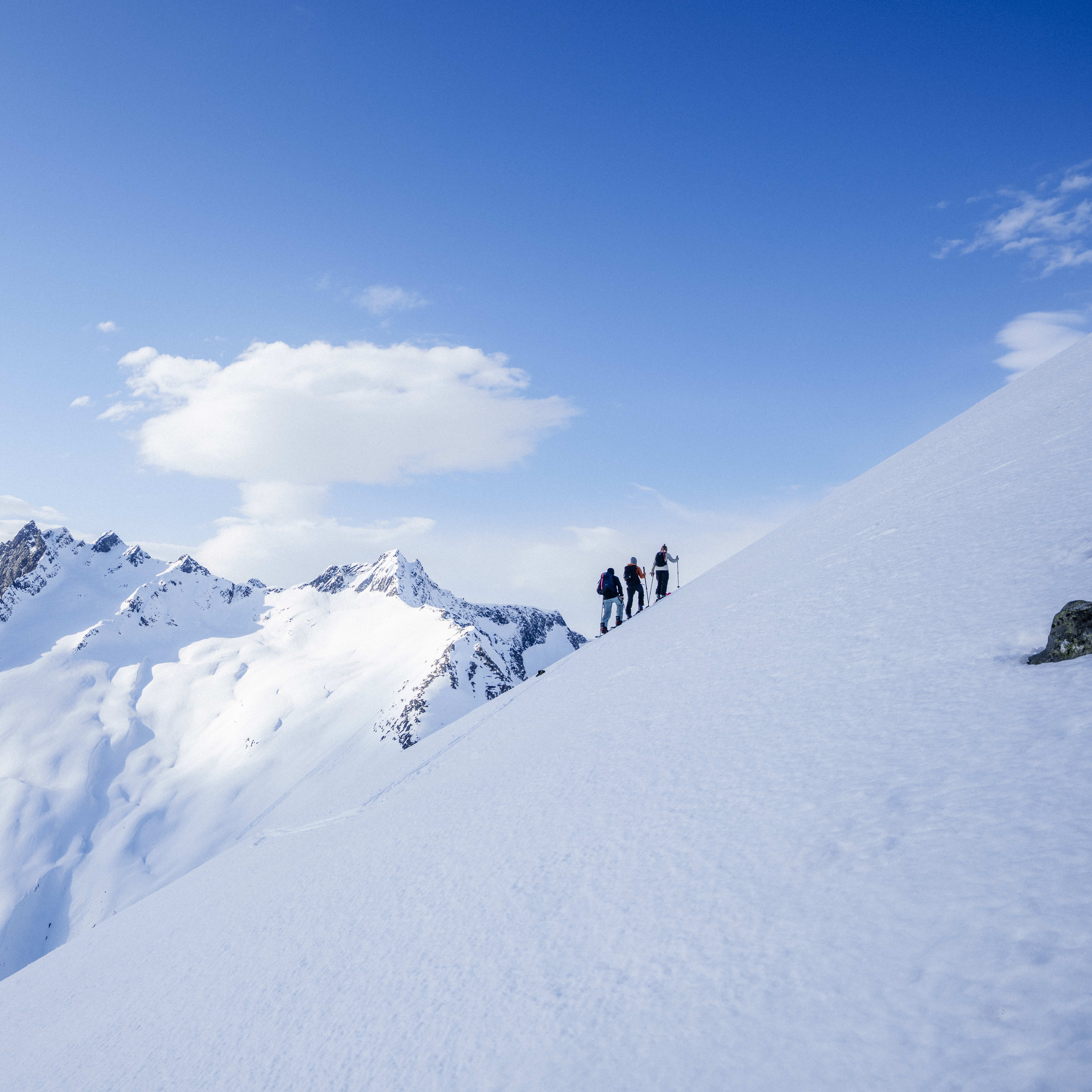 Three people hiking on a snowy mountain ridge. The sky is blue with white clouds.