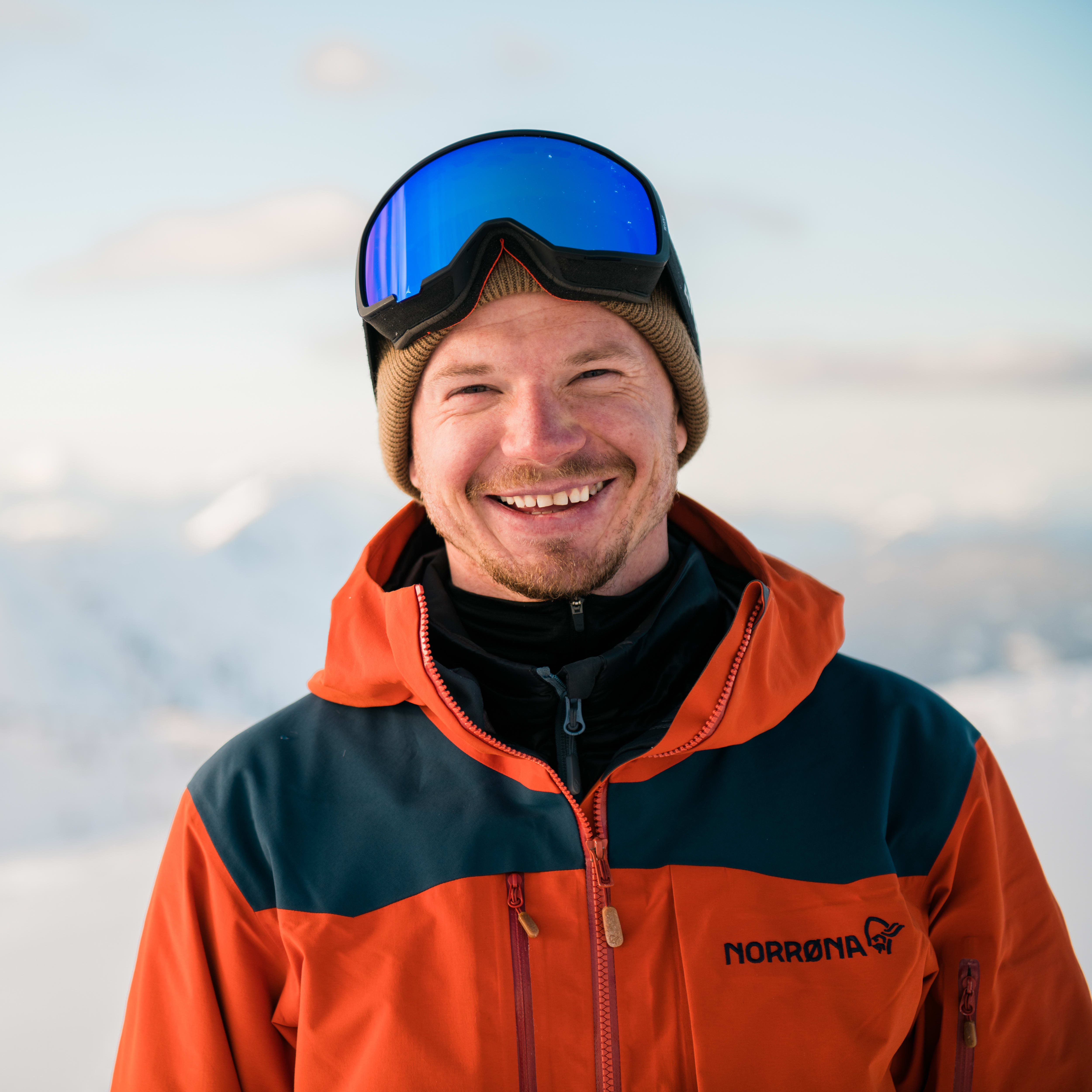 Smiling man wearing blue goggles, a black and orange Norrøna jacket, and a brown beanie, standing in front of a snow-covered mountain range. 
