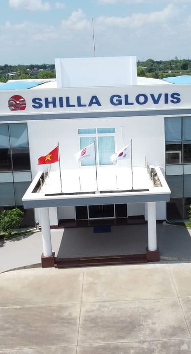 An aerial view of a large white building with the name "SHILLA GLOVIS" on the front. The building has large glass windows and a covered entrance with a flagpole displaying the flags of Vietnam, the United States, and South Korea.  The building is surrounded by landscaping and a paved parking lot. 
