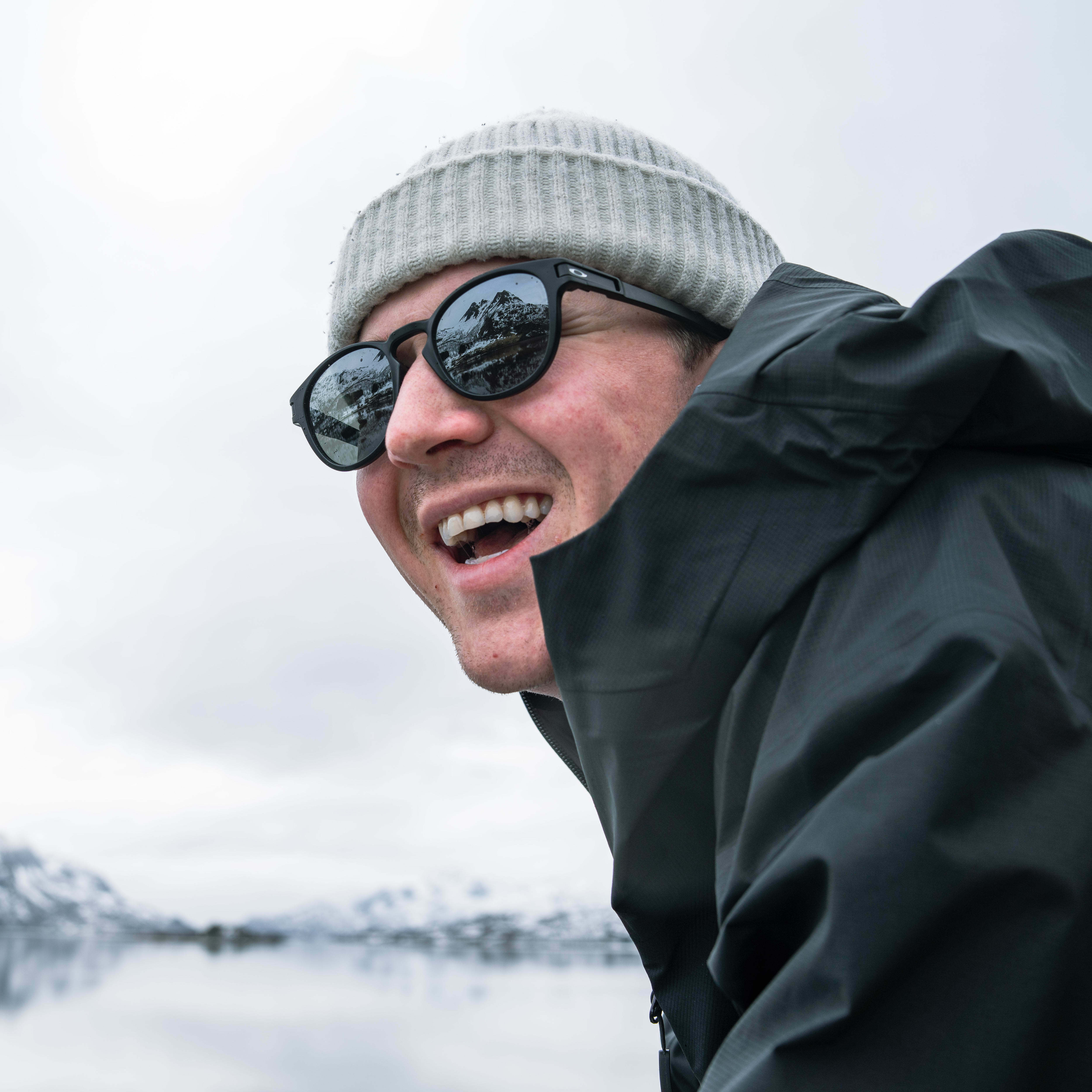 A man wearing a gray beanie and black sunglasses, is wearing a black Norrøna jacket. He is smiling and looking to the left. In the background, a blurry image of a body of water and snow covered mountains.