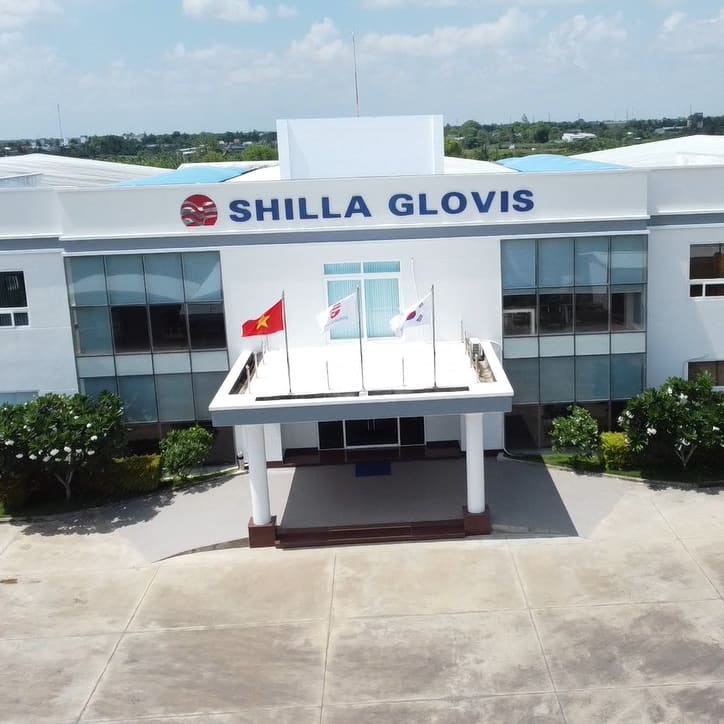 An aerial view of a large white building with the name "SHILLA GLOVIS" on the front. The building has large glass windows and a covered entrance with a flagpole displaying the flags of Vietnam, the United States, and South Korea.  The building is surrounded by landscaping and a paved parking lot. 
