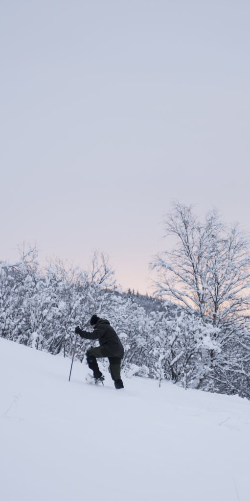 A person wearing Norrøna clothing is hiking up a snowy mountainside.  The sky in the background is a pale pink.  