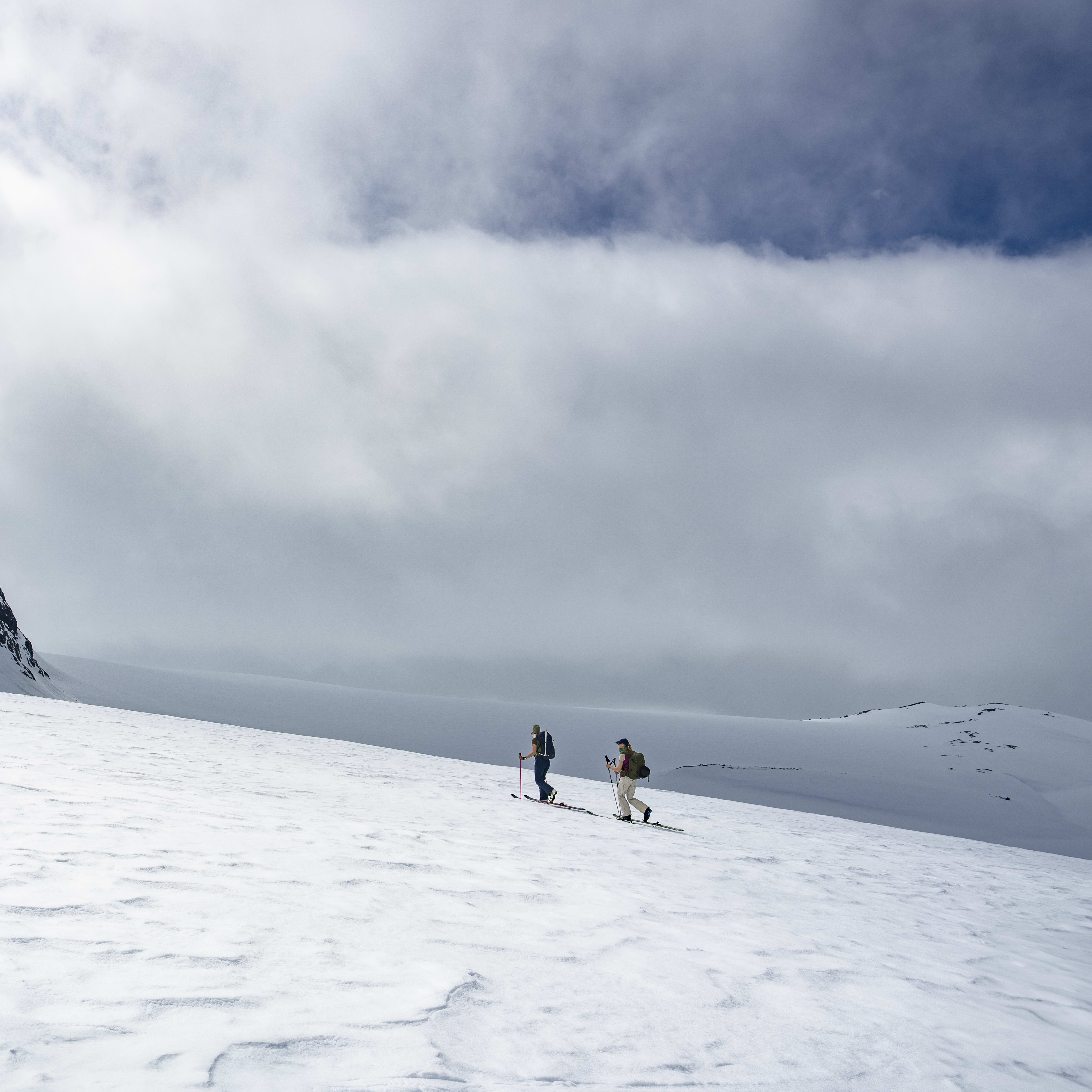 Two people ski across a snowy mountain landscape wearing Norrøna gear.  The sky is cloudy. 
