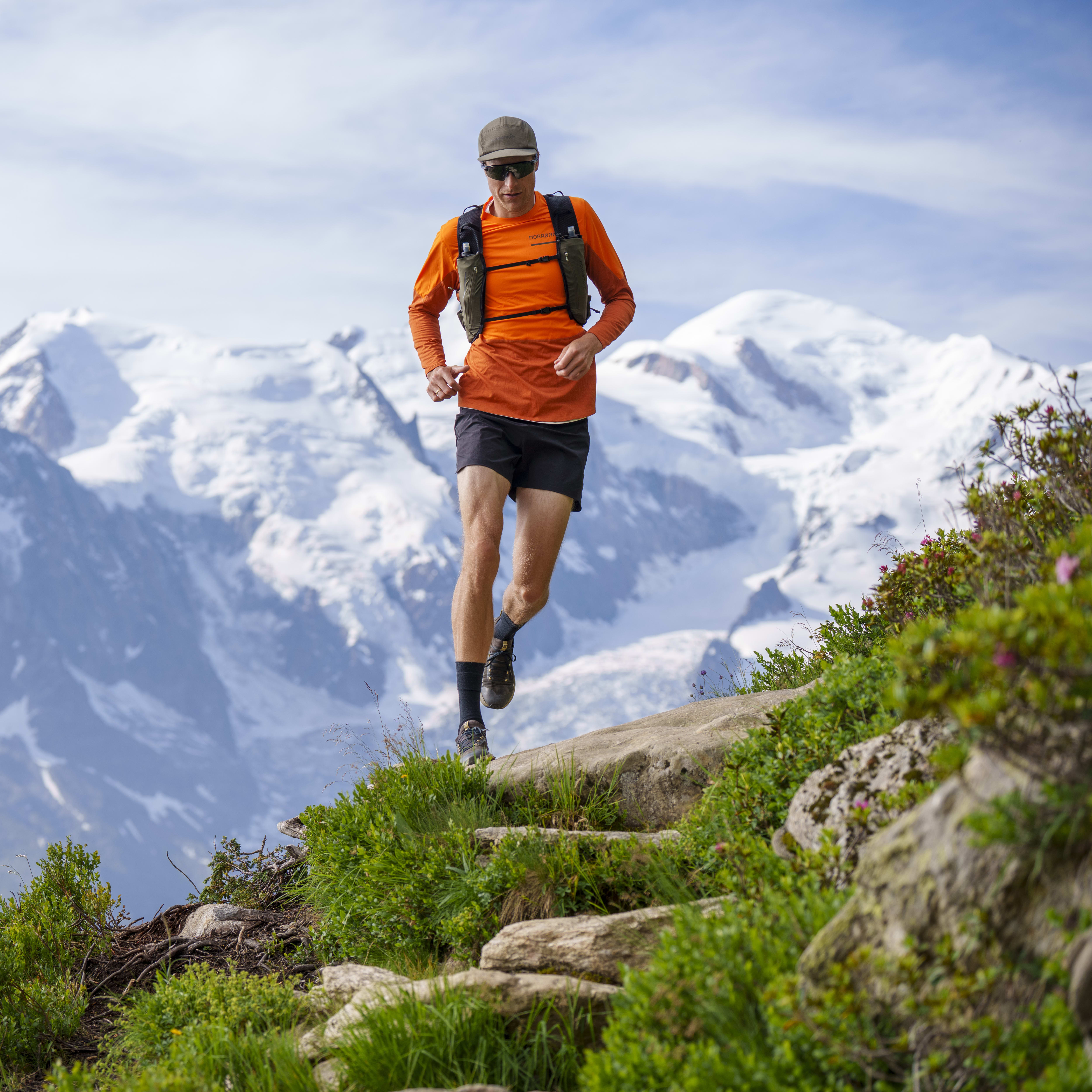 A man trail runs downhill. He wears an orange Norrøna shirt and carries a pack. Snowy mountain peaks are in the background.