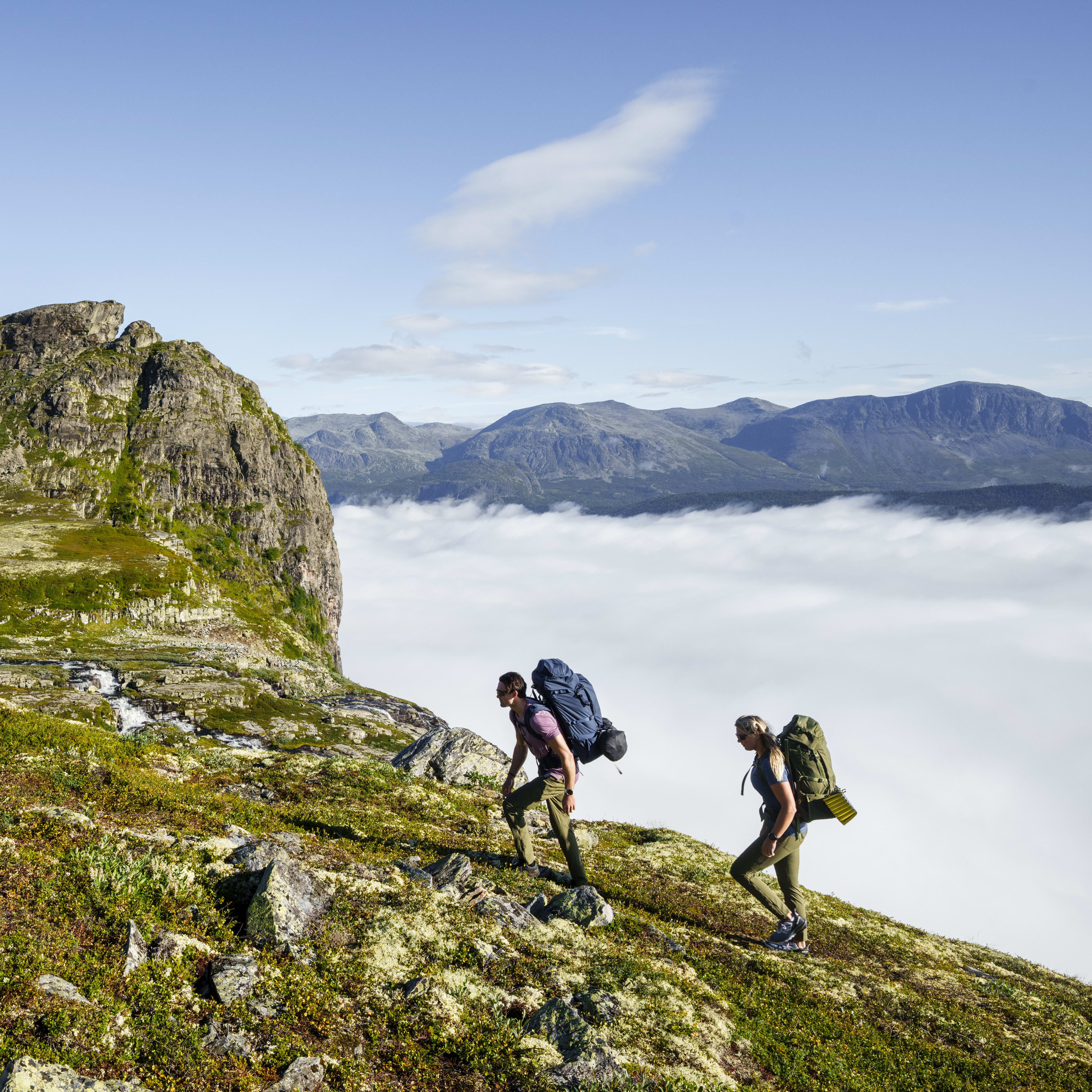 Two people hike up a rocky mountainside with backpacks, wearing Norrøna clothing, towards a foggy valley.