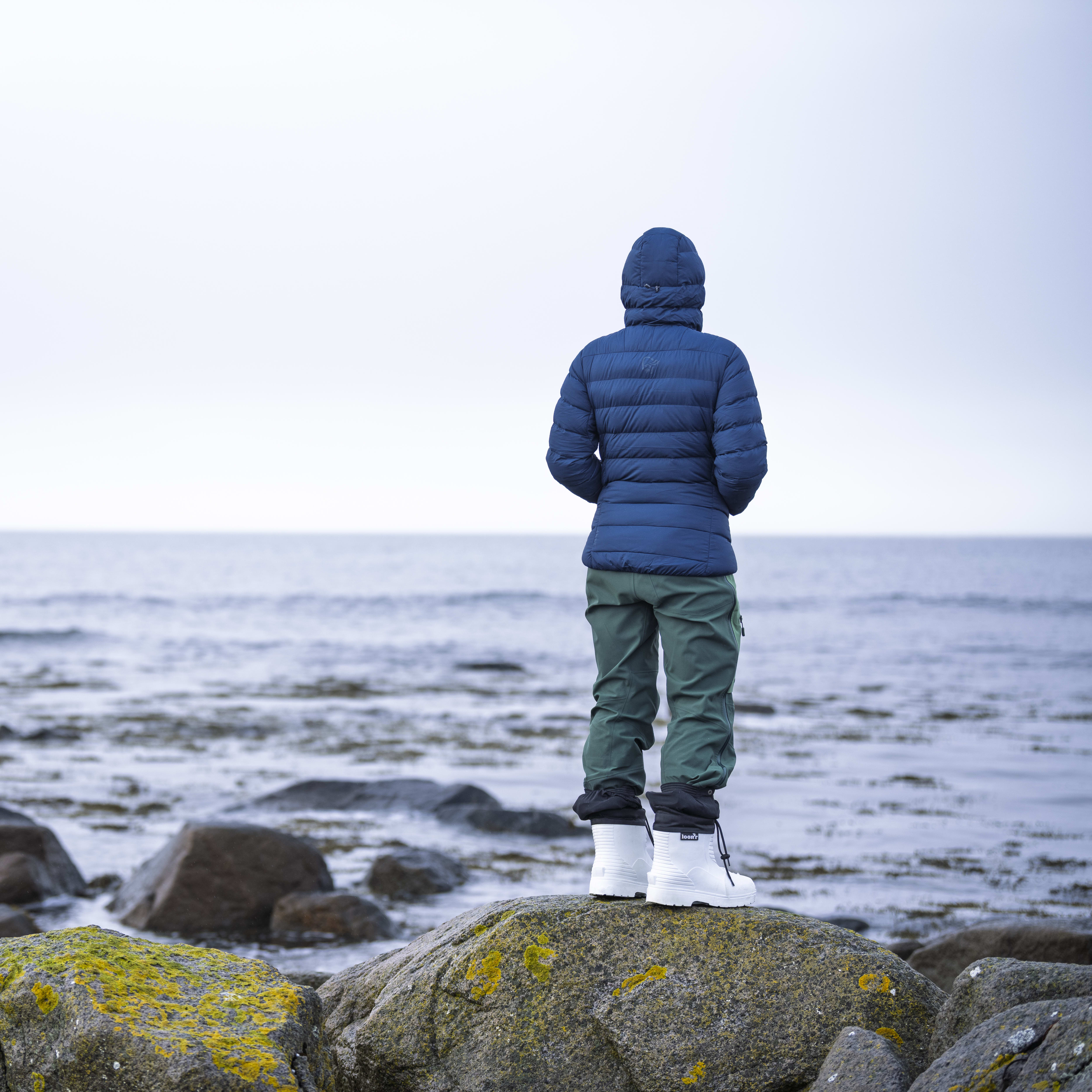 Person wearing a Norrøna jacket and white boots standing on rocks by the sea.