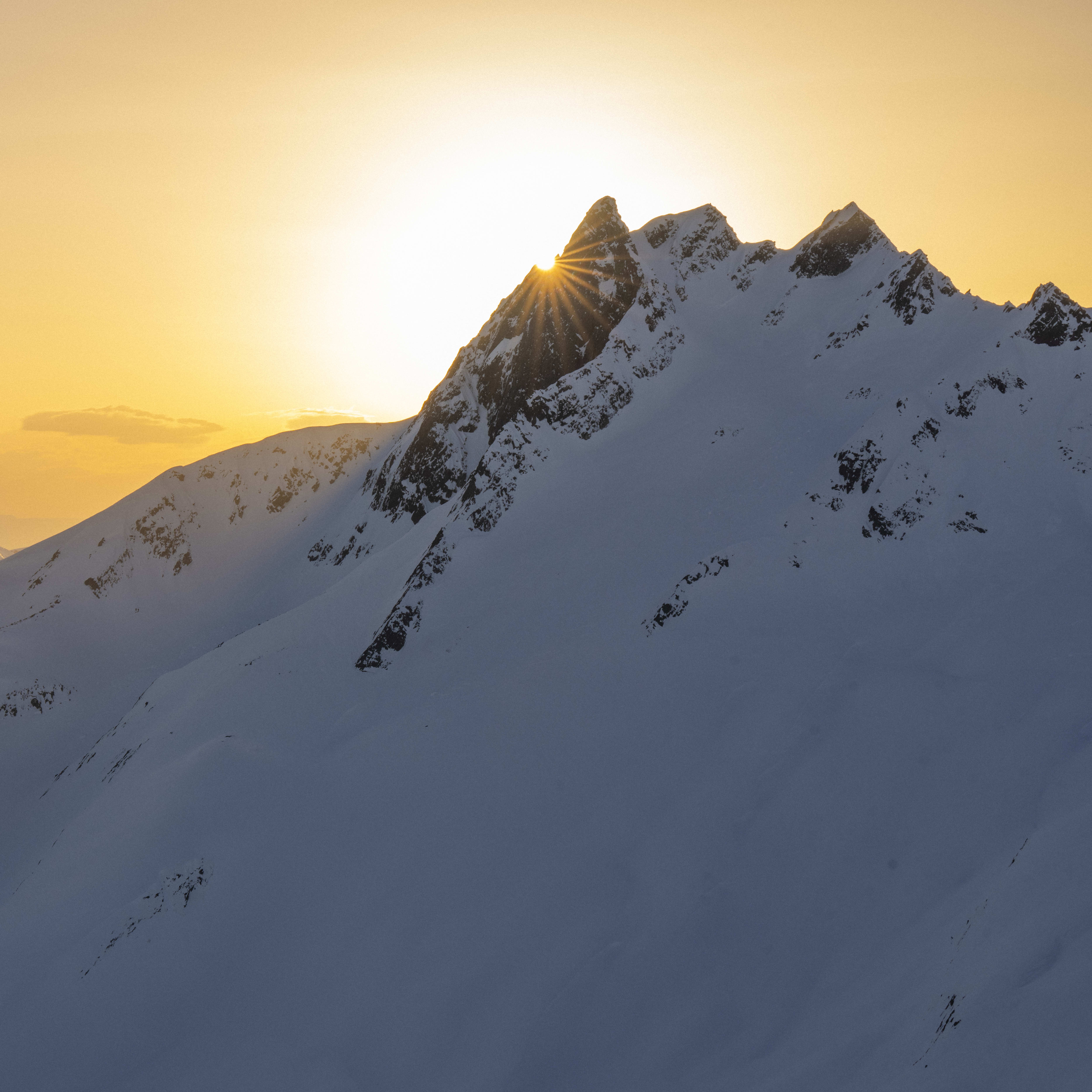 A view of snow-capped mountains at sunset with the sun shining through a peak.