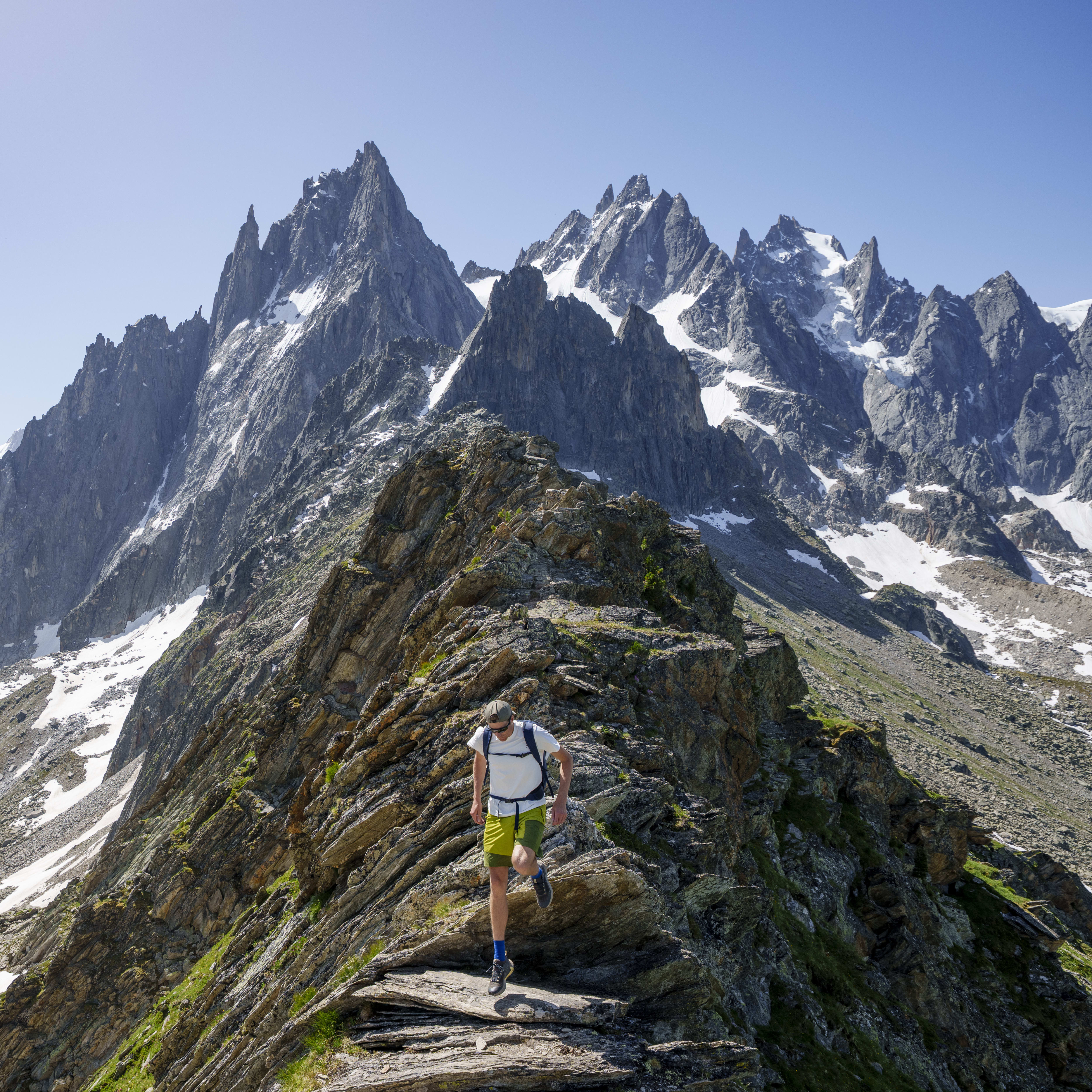 A man hikes on a rocky mountain trail with snowy peaks in the background. He wears Norrøna shorts and a white t-shirt with a backpack.