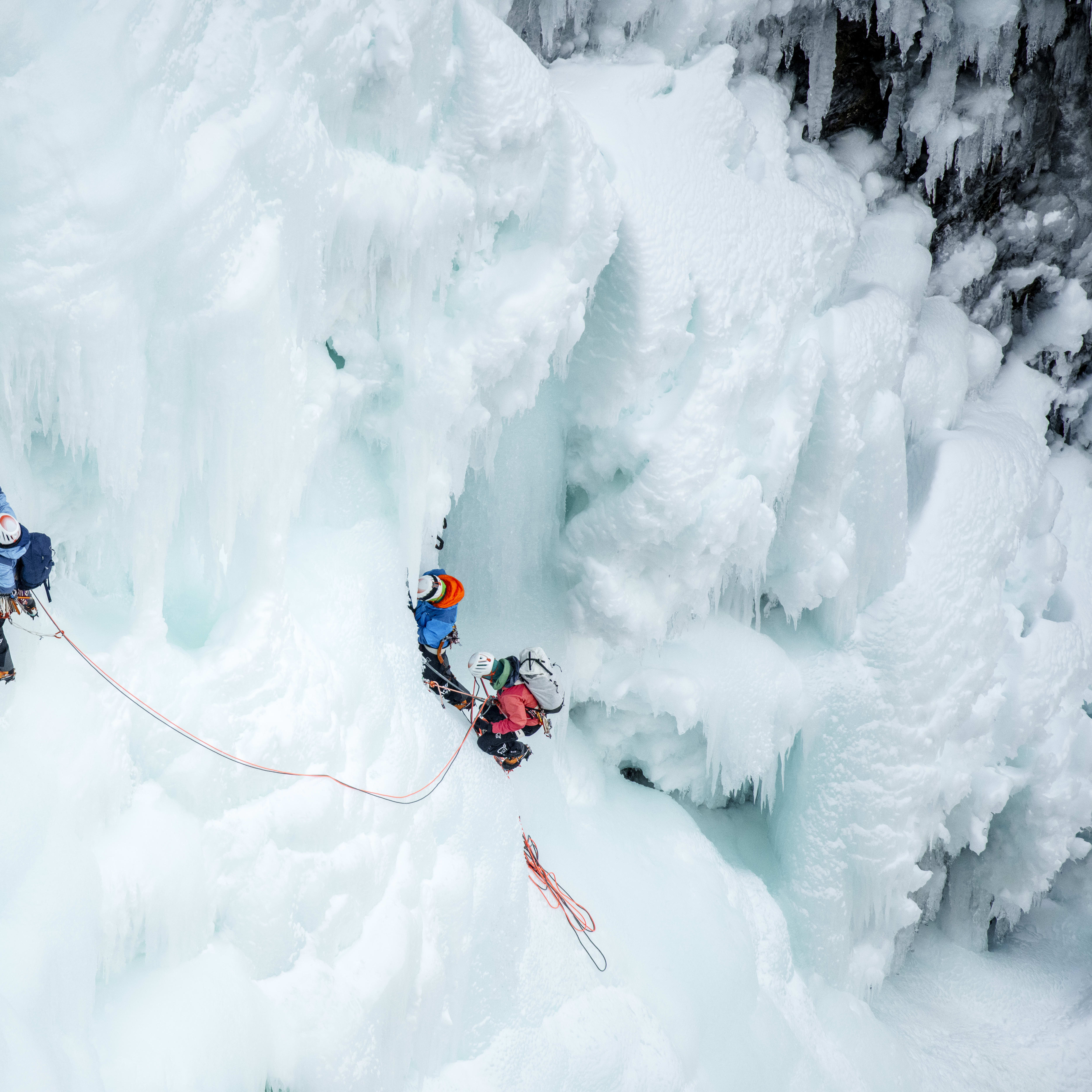 Two ice climbers ascend a frozen waterfall, one wearing a blue Norrøna jacket, secured by ropes on a bright, icy surface.

