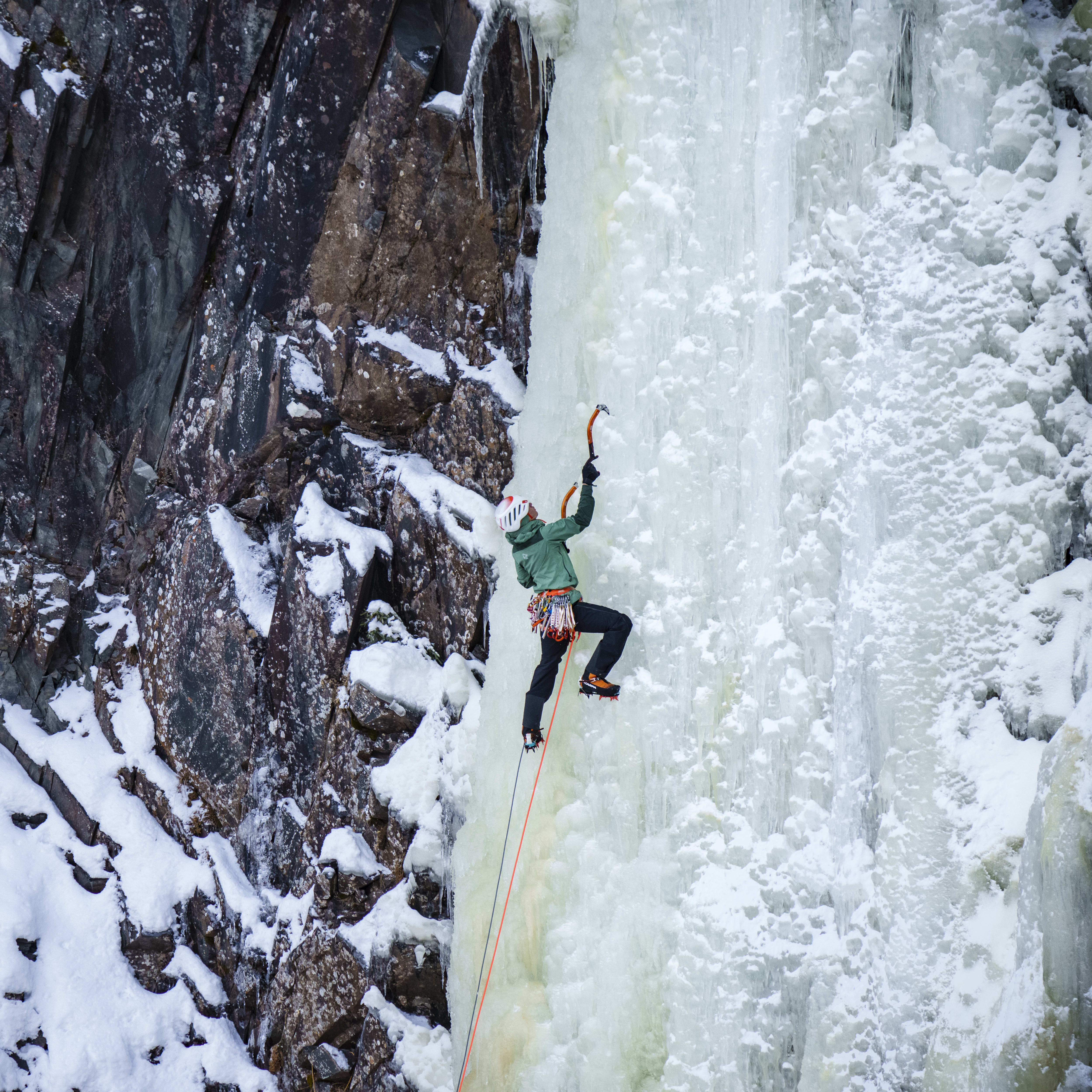 A person ice climbing wearing a green Norrøna jacket and black pants. The climber is attached to a rope and uses ice axes to ascend a frozen waterfall.