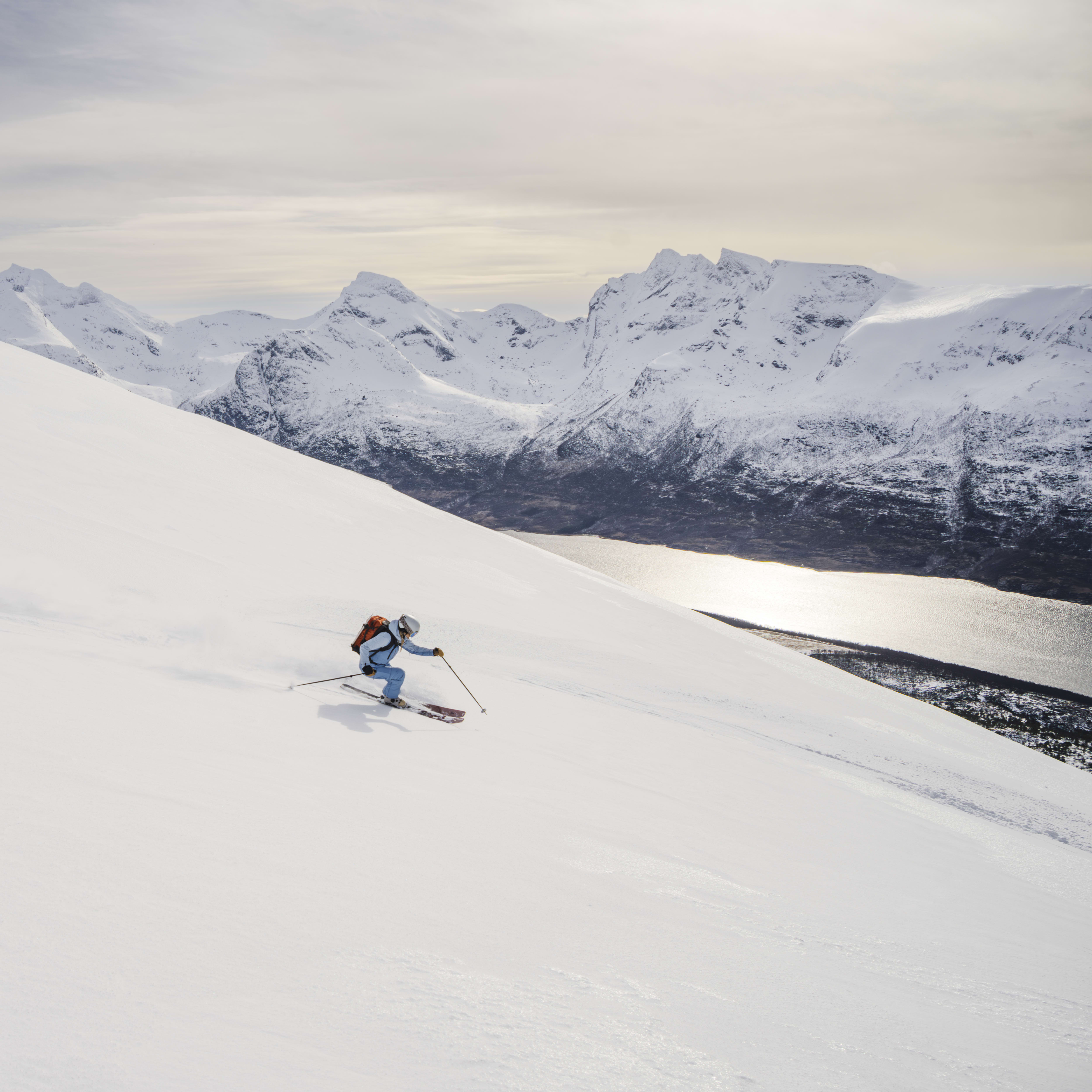 A skier wearing Norrøna outerwear skis down a snowy slope with mountains and a body of water in the background.
