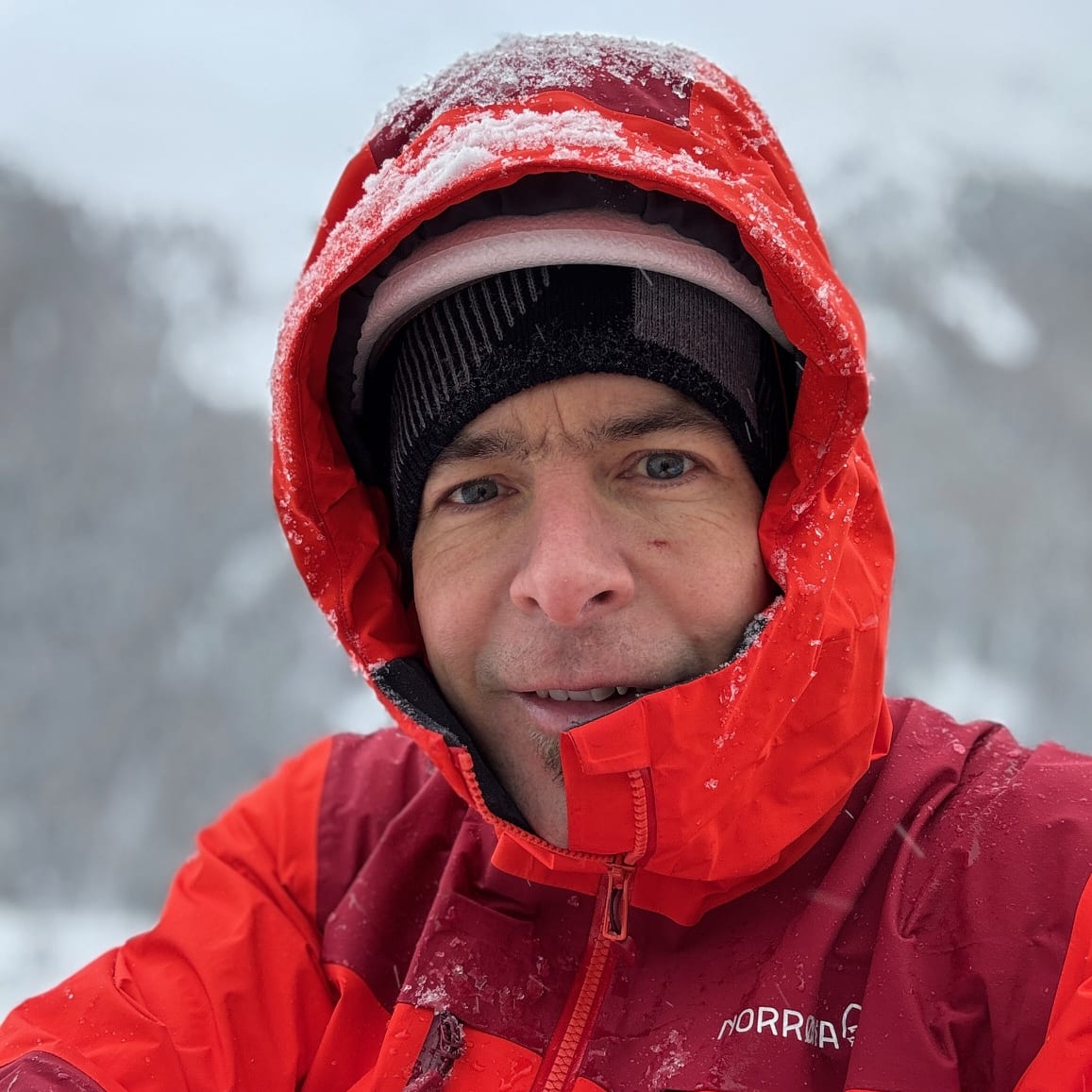 A man in a red Norrøna jacket with a black hat, looking directly at the camera. Snow falls around him. 
