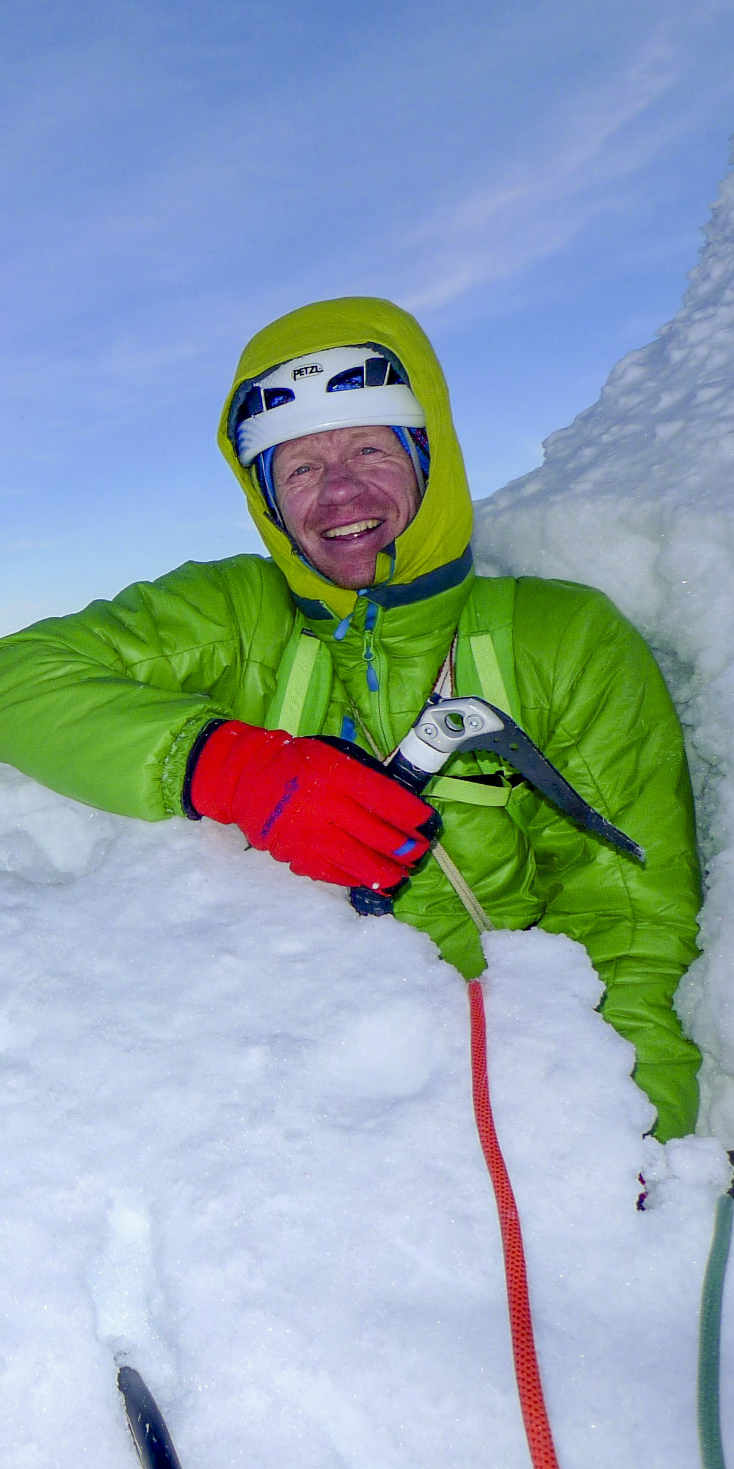 A man wearing a green jacket, red gloves, and a helmet is smiling at the camera. He is standing in the snow on a mountain peak, with a large rocky mountain in the background.  Two ice axes and a red rope are in the snow around the man.