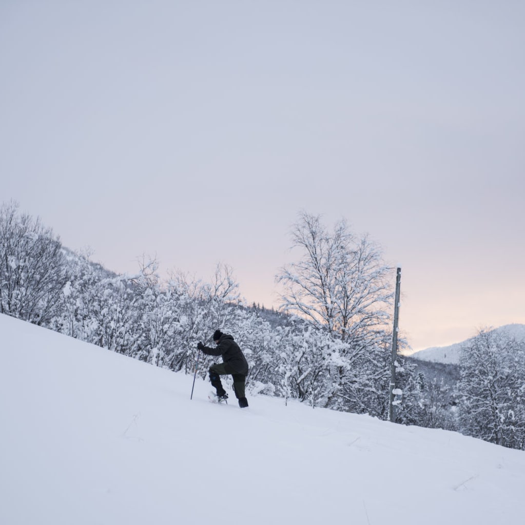 A person wearing Norrøna clothing is hiking up a snowy mountainside. The sky in the background is a pale pink.