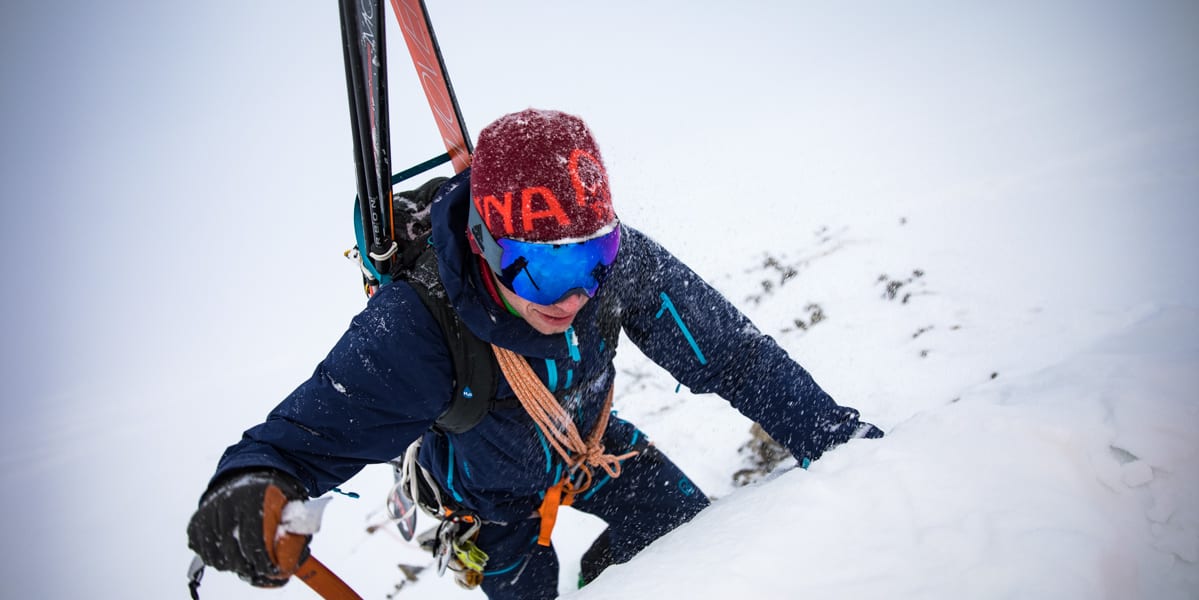 A climber ascending a snowy slope, wearing a Norrøna jacket and beanie, goggles, and carrying skis. He uses an ice axe.
