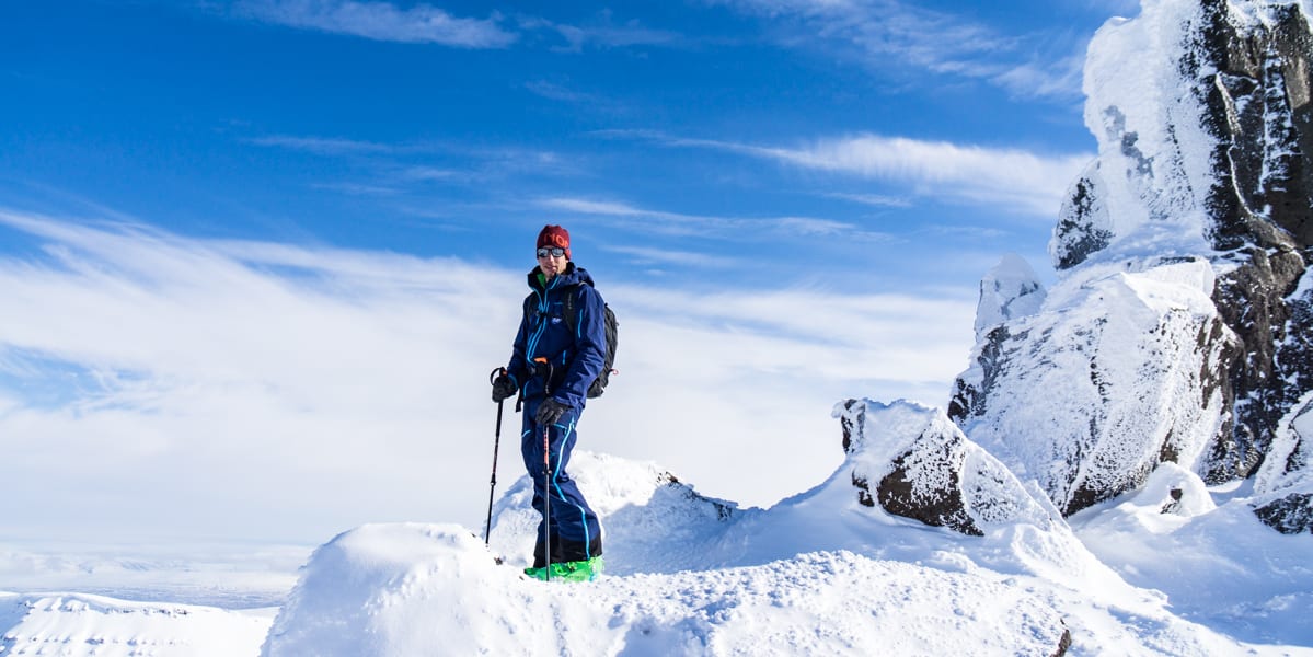 A person stands on a snow-covered mountain wearing a Norrøna jacket and pants, using hiking poles, with a clear blue sky overhead.

