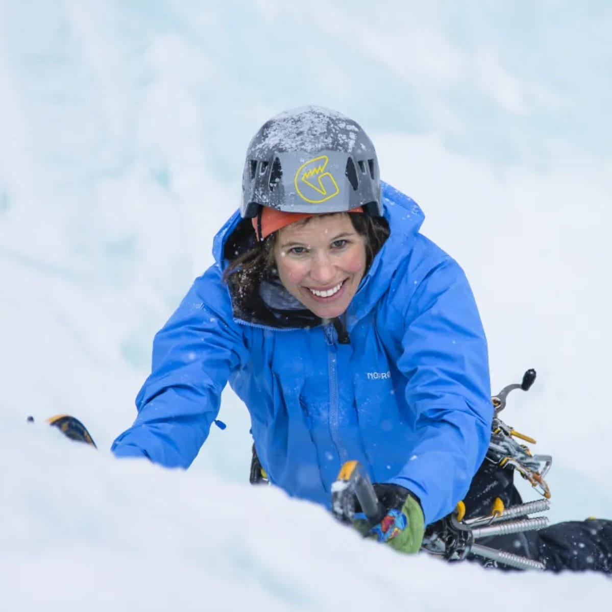 Smiling woman ice climbing, wearing a helmet and blue Norrøna jacket. Ice axes and gear visible against the snowy backdrop.