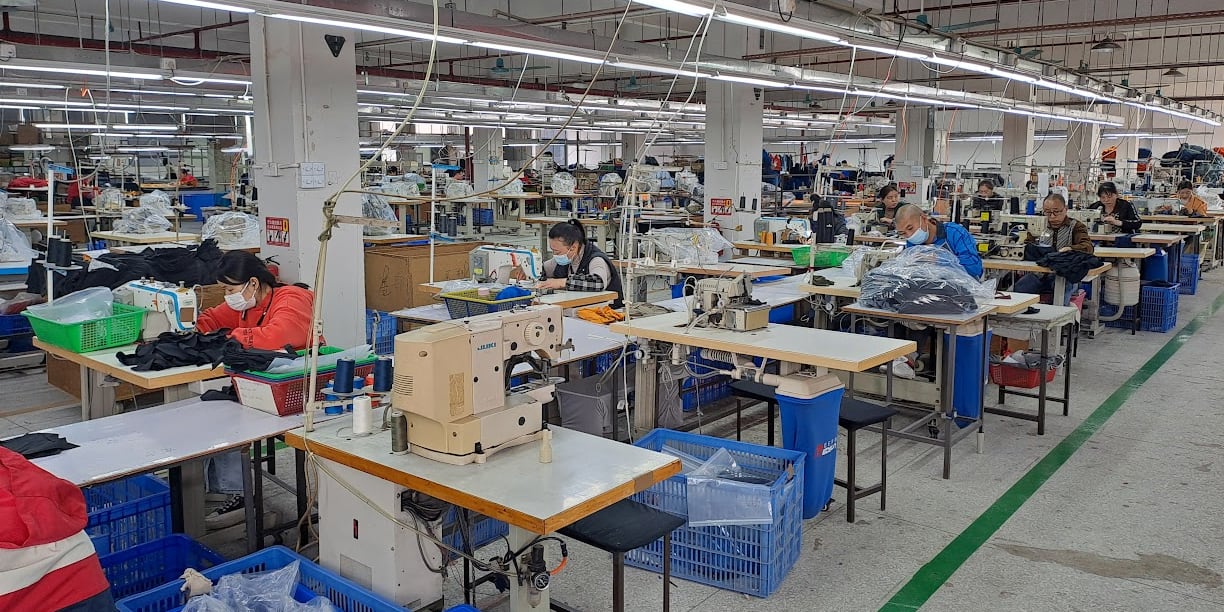 A factory floor with several rows of sewing machines. Workers are seated at the machines, sewing Norrøna clothing.  There are blue plastic bins and black stools throughout the space.  The floor has a green stripe running along the side of the image. 

