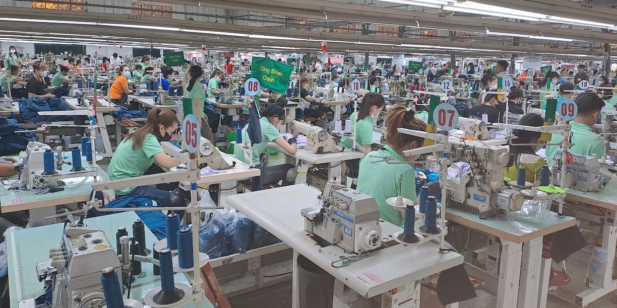 A wide shot of a sewing factory with many workers, all wearing face masks, operating sewing machines.  The workers are all wearing Norrøna clothes. 
