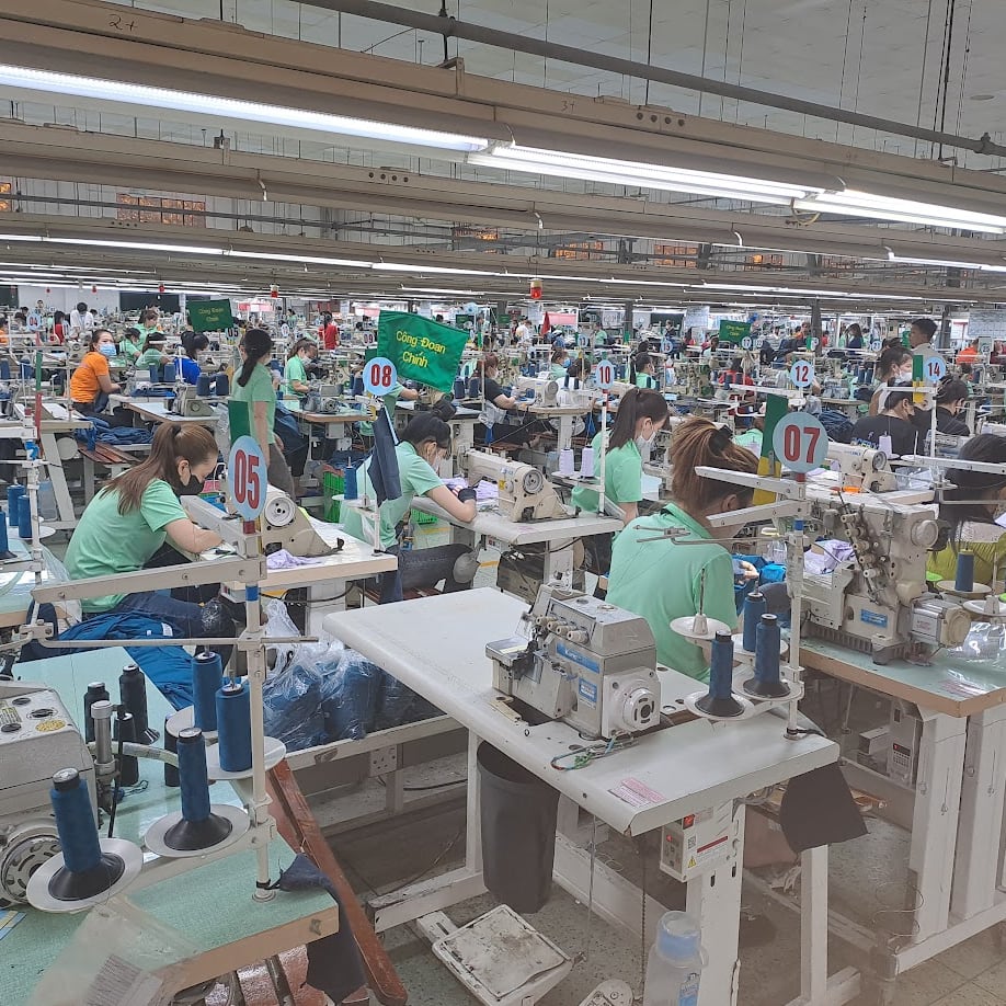 A wide shot of a sewing factory with many workers, all wearing face masks, operating sewing machines. The workers are all wearing Norrøna clothes.