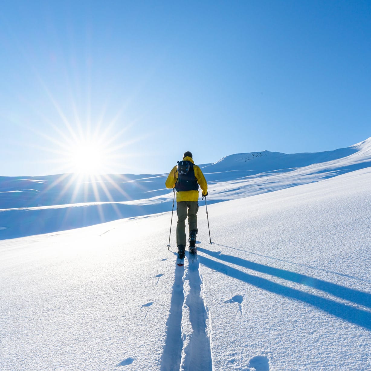 A person in a yellow jacket and backpack is backcountry skiing on a snowy, bright, sunny day. The sun is shining brightly in the sky, and the person is leaving a track in the snow.
