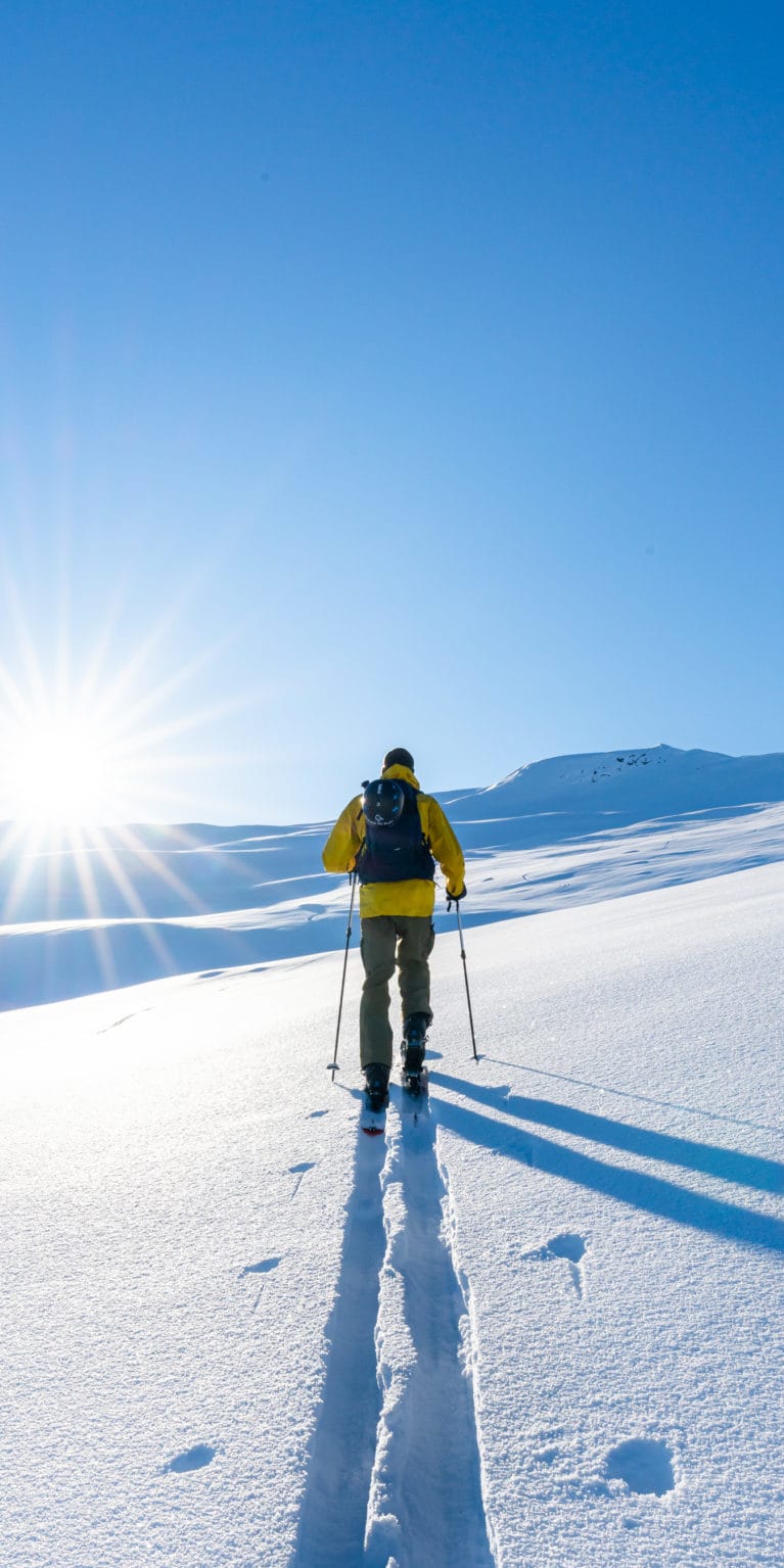 A person in a yellow jacket and backpack is backcountry skiing on a snowy, bright, sunny day. The sun is shining brightly in the sky, and the person is leaving a track in the snow.  