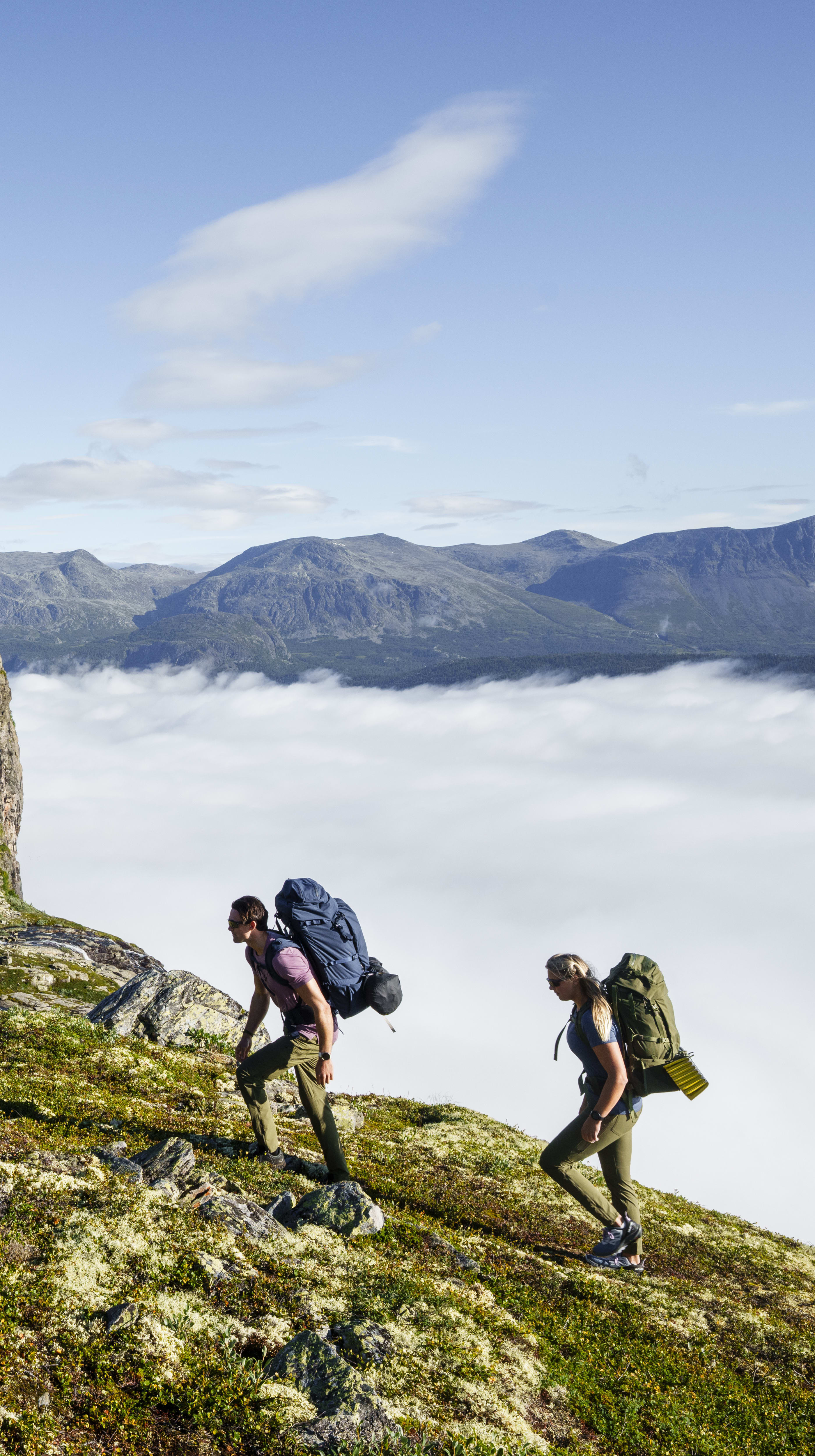 Two people hike up a rocky mountainside with backpacks, wearing Norrøna clothing, towards a foggy valley. 
