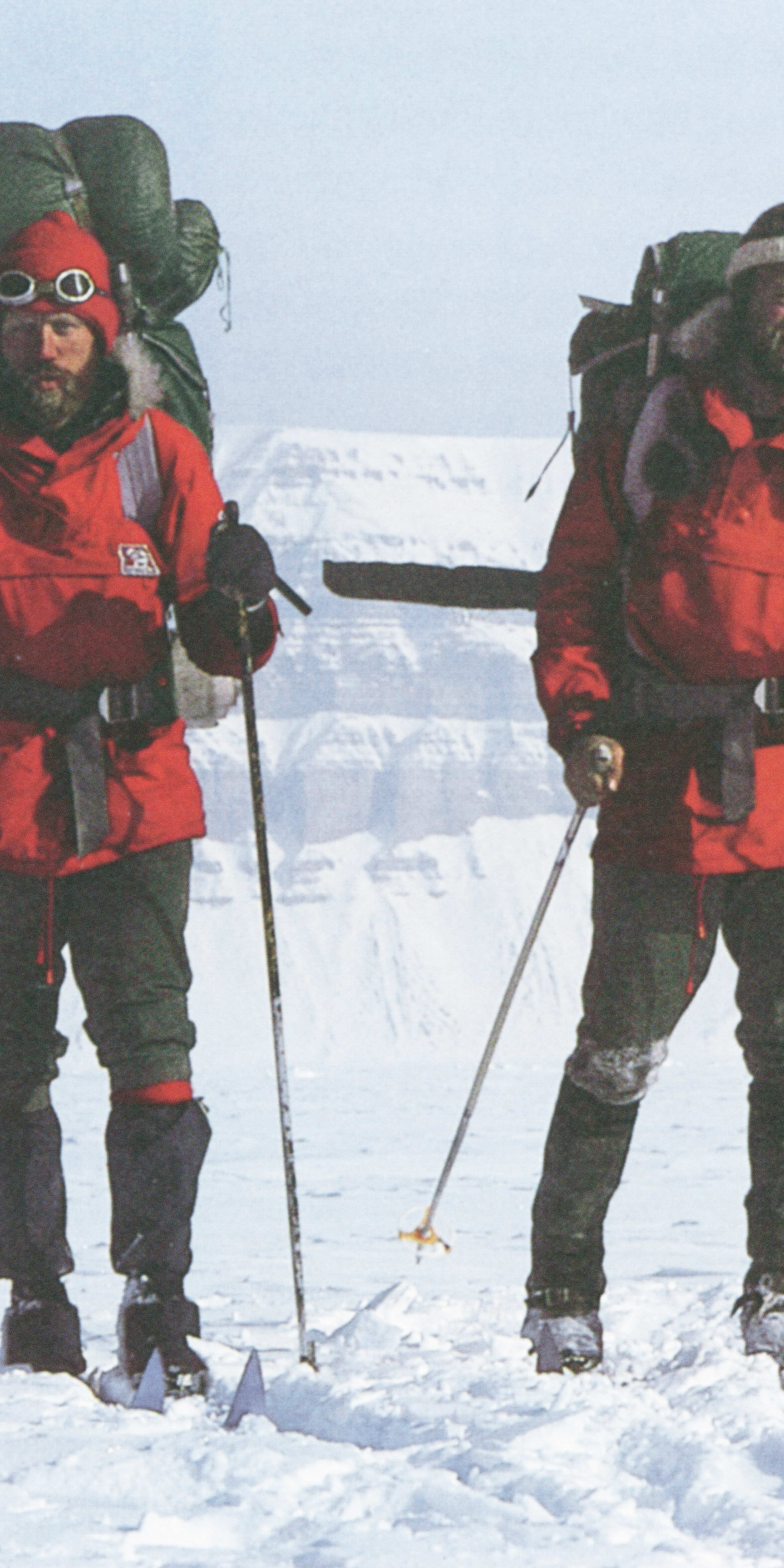 Two men are cross-country skiing in a snowy, mountainous landscape. Both men are wearing red jackets, backpacks, and ski poles.  The sky is a light blue.