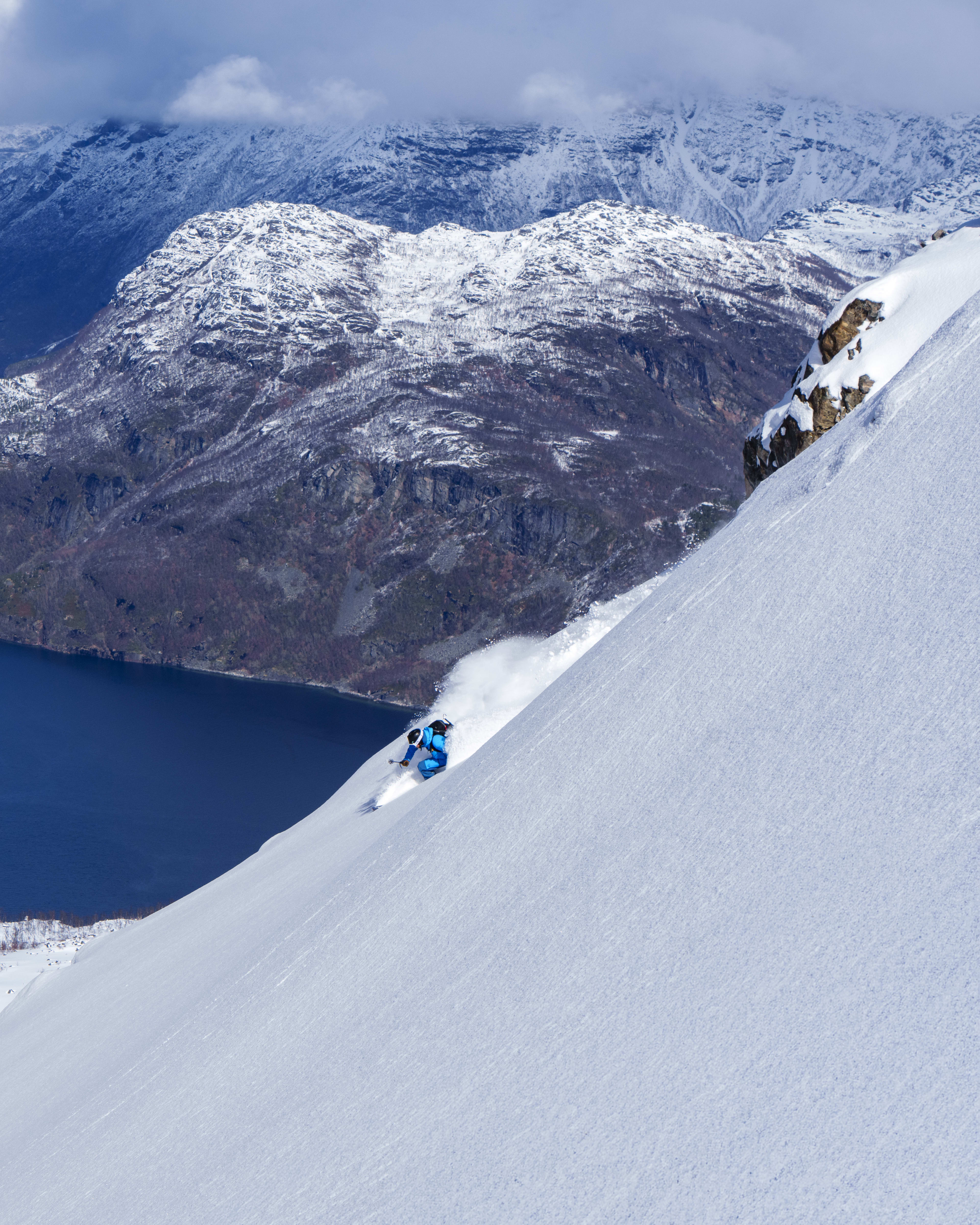 A skier in blue Norrøna gear skis down a steep snowy slope with a blue fjord and mountains in the background.