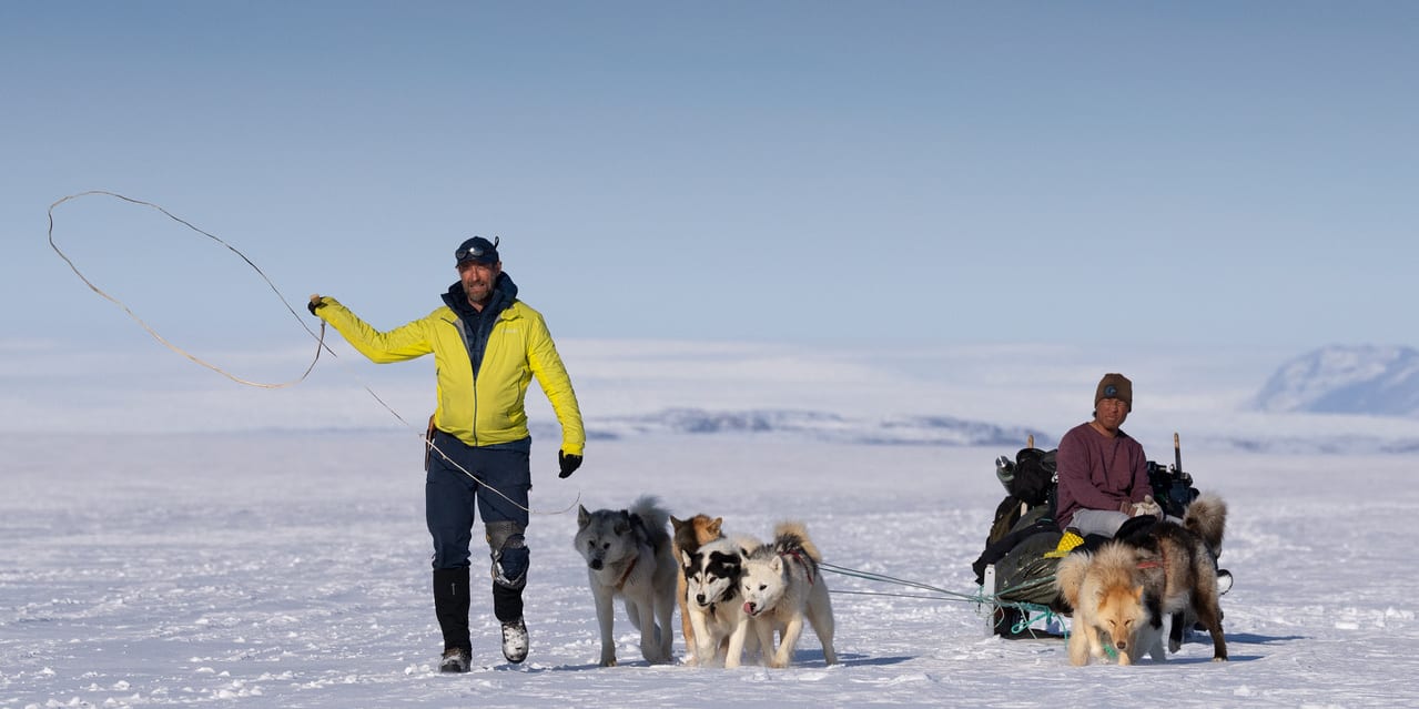 Two people in winter gear, one running with a rope, the other sitting on a sled, with a team of dogs pulling the sled.  
